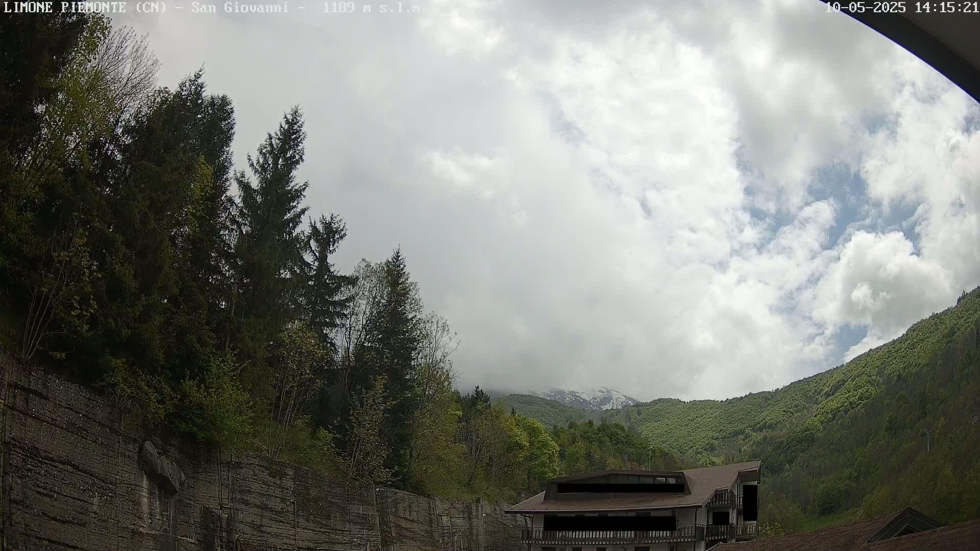 A cloudy day reveals a mountainside covered in lush green trees and a partially visible building, with a rocky wall and more trees in the foreground.