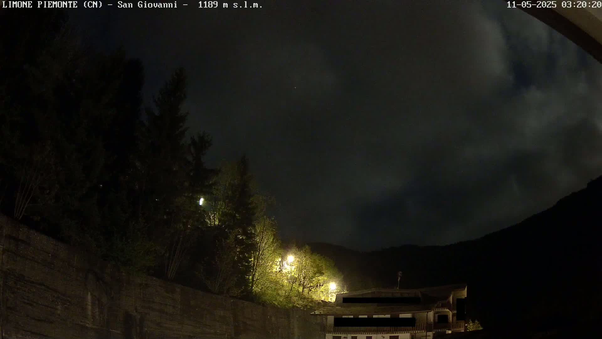 A nighttime view of a dark, cloudy sky over a hillside with trees and a building, illuminated by a few lights.