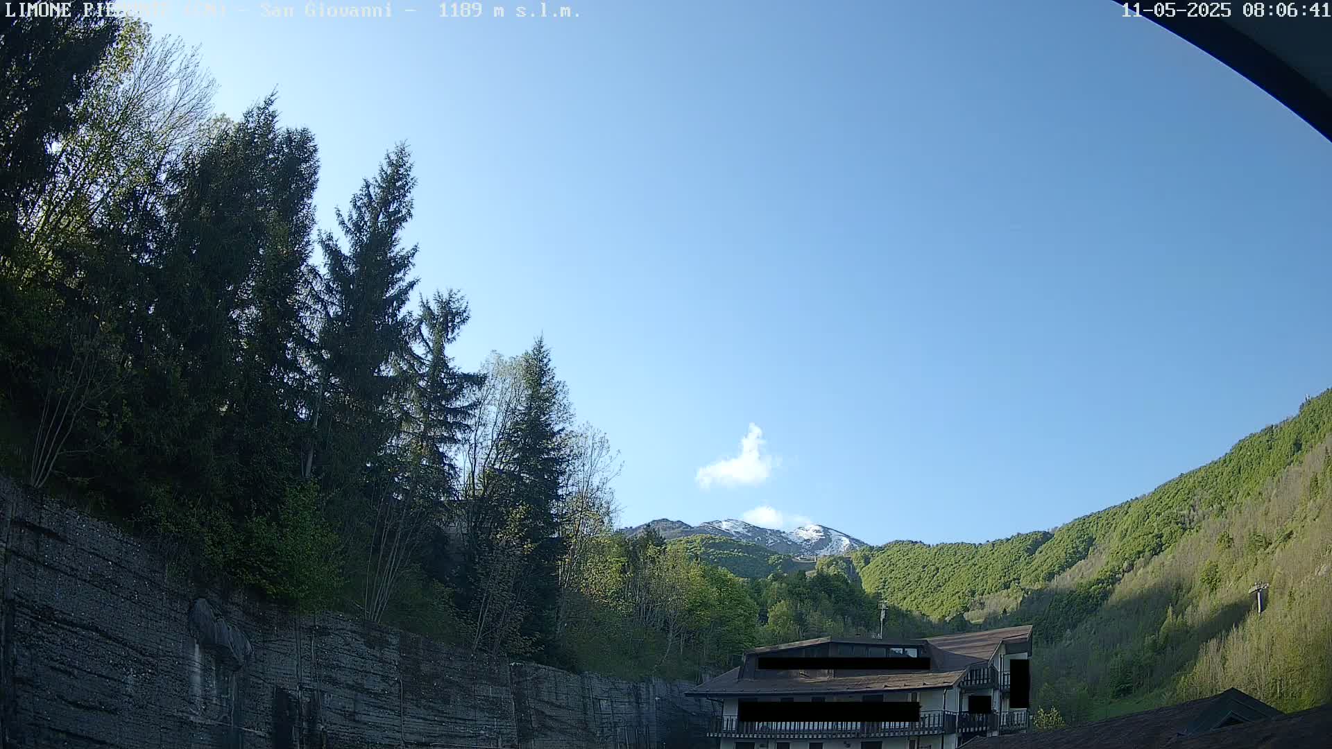 A mountainside with green trees and a building is visible under a clear blue sky, with a snow-capped mountain peak in the distance.