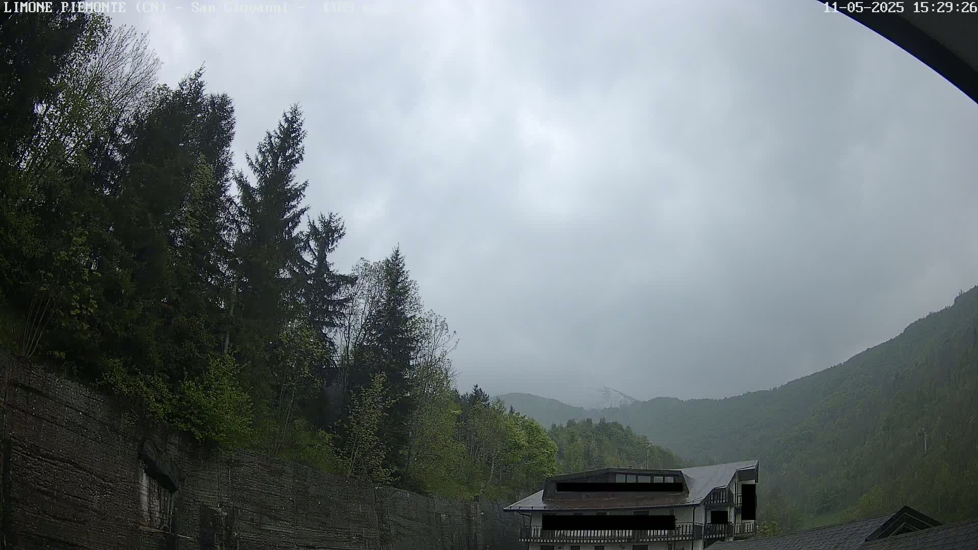 A cloudy day reveals a hillside dotted with evergreens and a building nestled amongst the trees, with mountains visible in the distance.