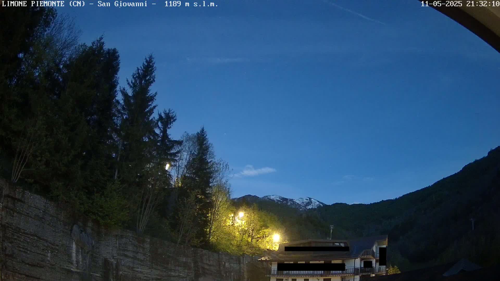 Under a twilight blue sky, dark evergreen trees and a building with lights illuminate a mountainside at night.