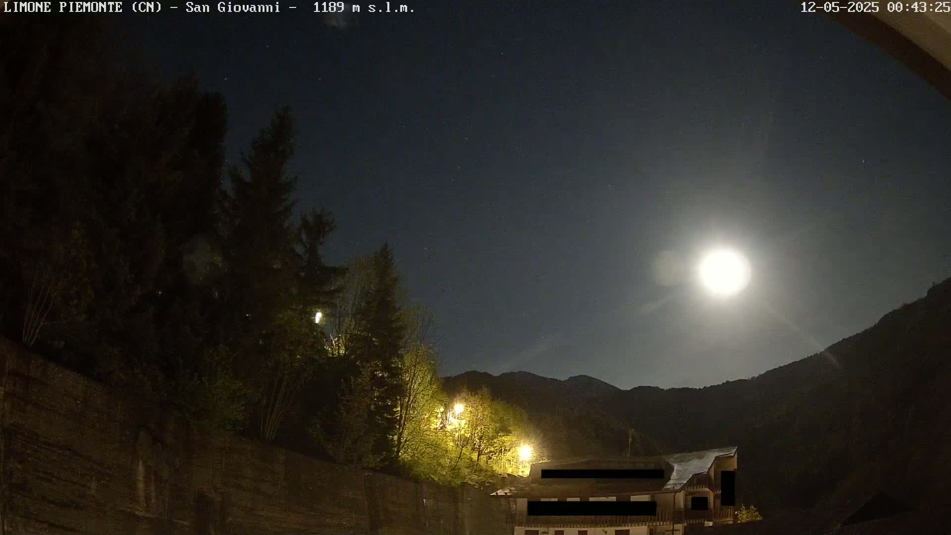 A bright moon illuminates a nighttime scene of trees, a building, and mountains under a clear sky.