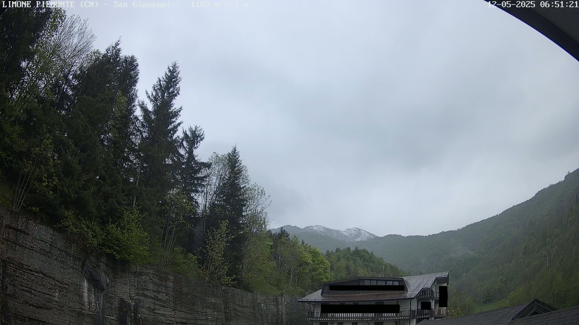 A cloudy day reveals a mountain range with a dusting of snow in the distance, partially obscured by lush green trees and a building nestled among them near a rocky cliff.