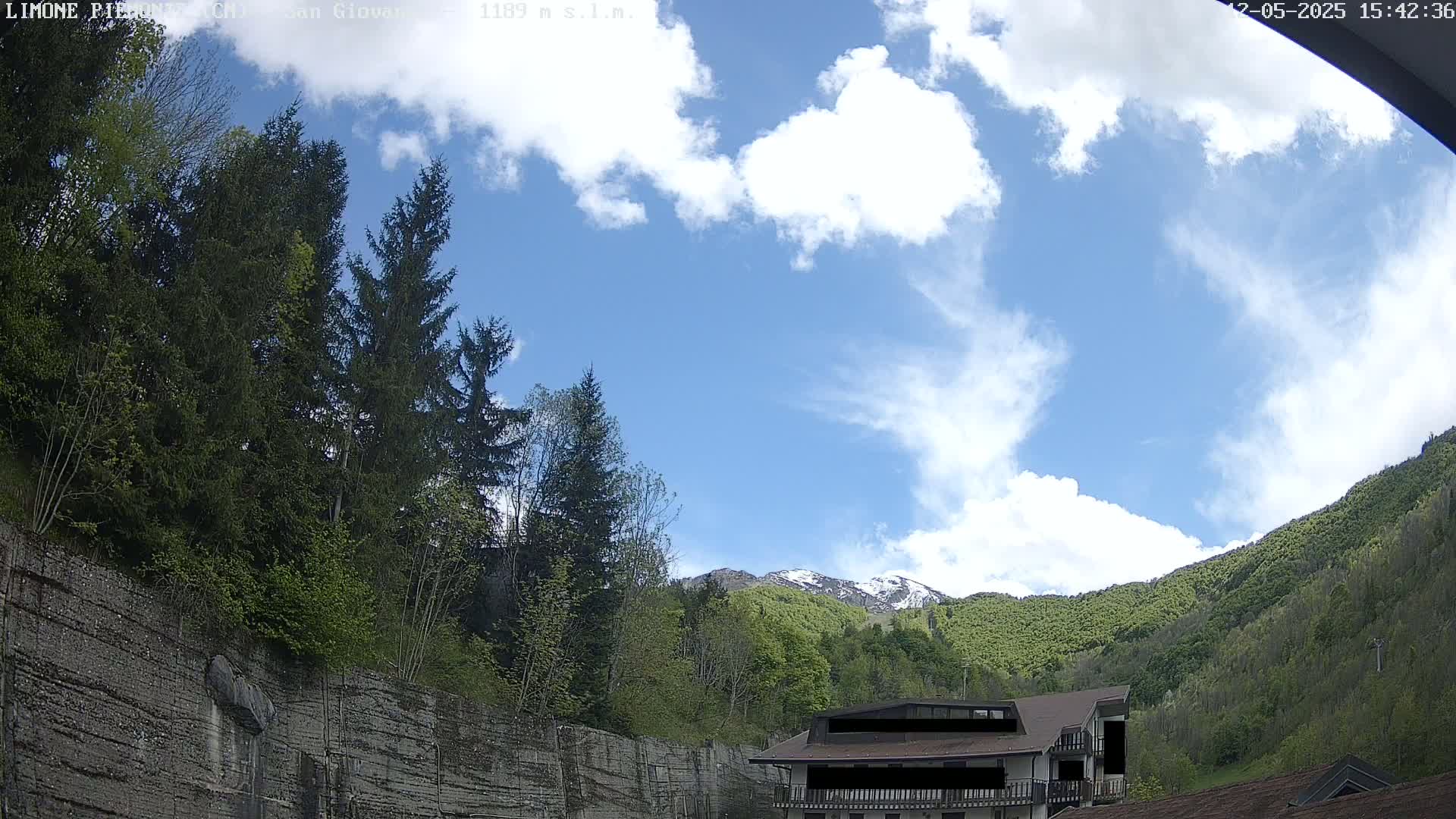 A partly cloudy blue sky overlooks a mountainous landscape with green trees, a building, and a stone wall.