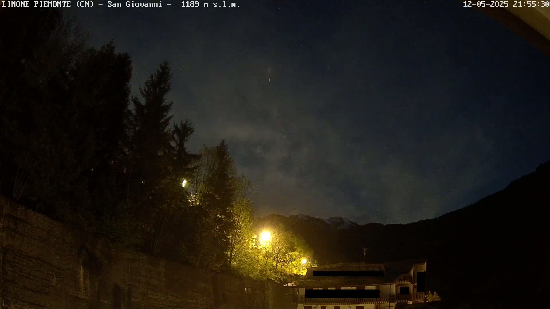 A nighttime view shows dark trees and buildings illuminated by streetlights against a mostly clear, starry sky with a mountain in the background.