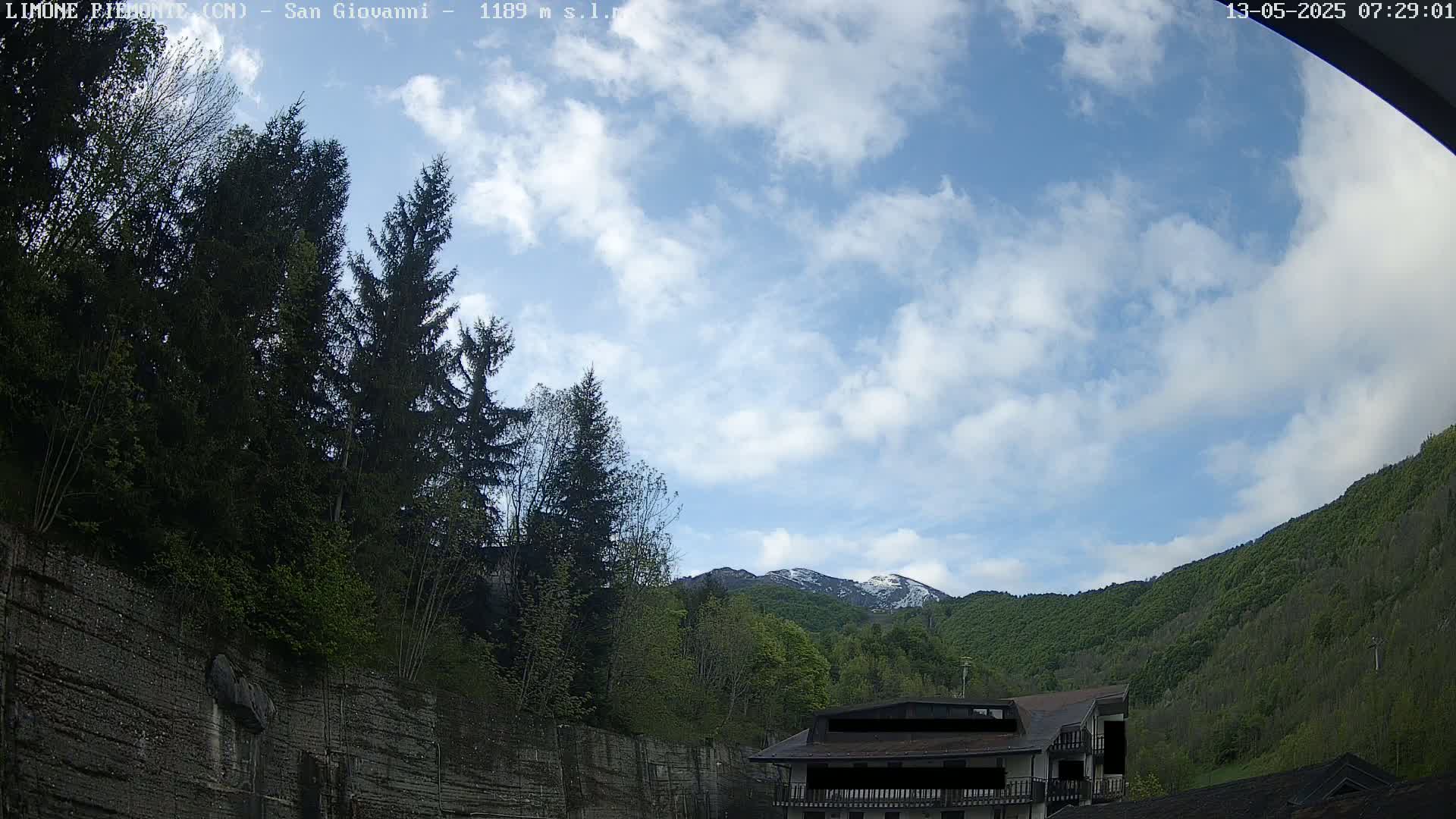A partly cloudy sky overlooks a mountain range with some snow, green trees, and a building nestled among the greenery.