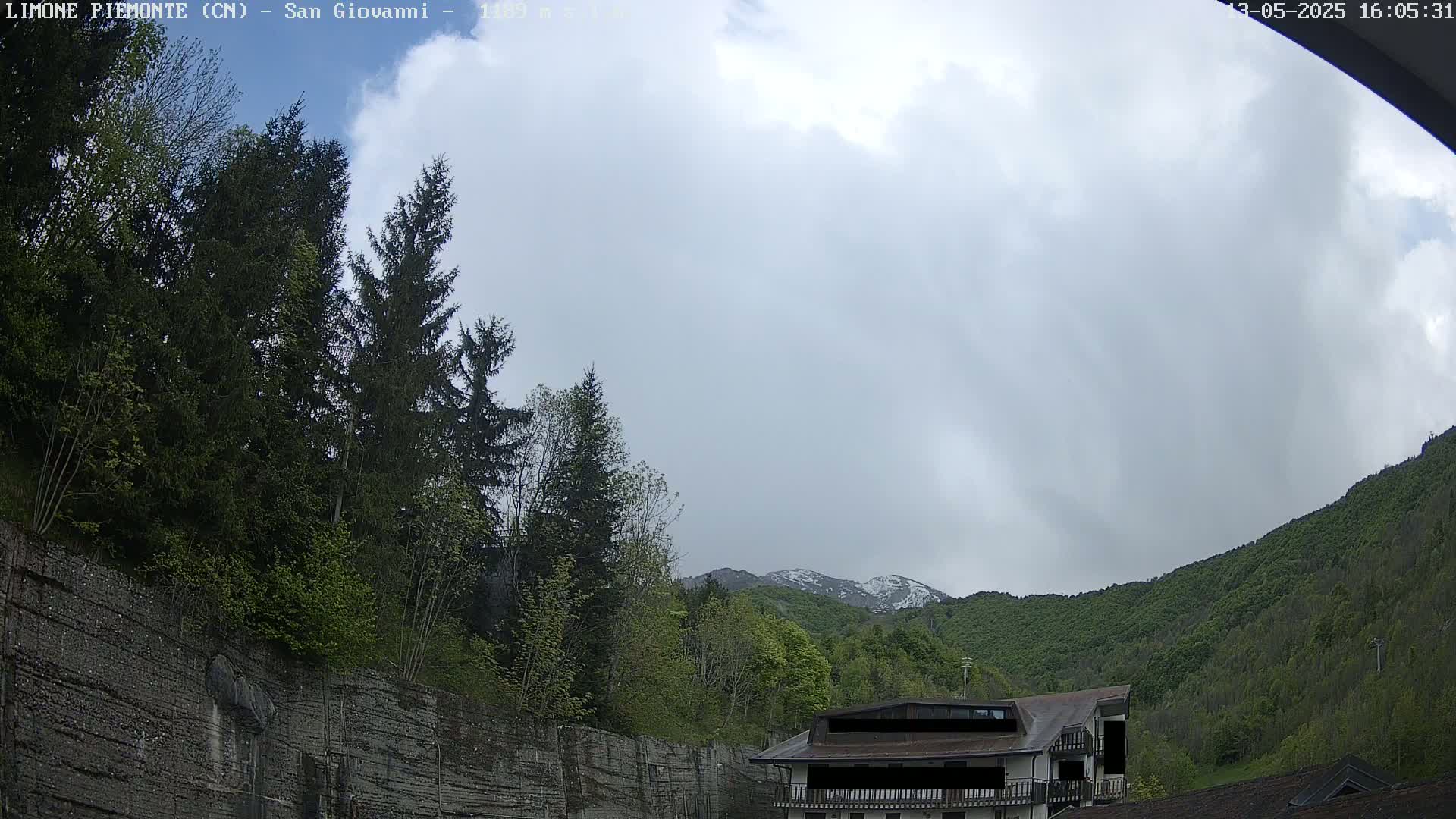 A cloudy day reveals a mountain range partially covered in snow, visible behind a building and a wall of trees and vegetation.