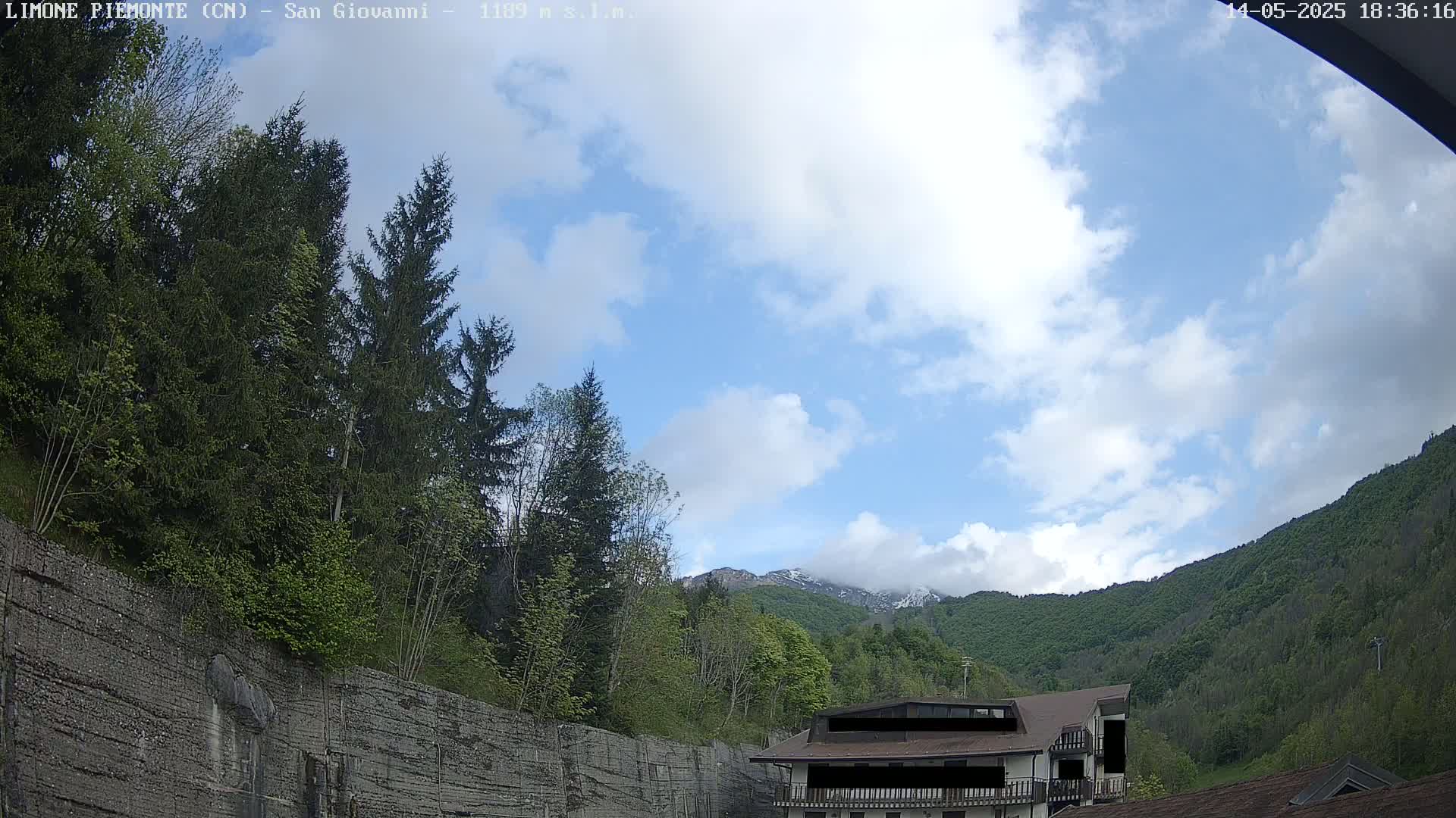 A partly cloudy sky overlooks a mountainside covered in lush green trees and a building nestled at the base of a rocky embankment.