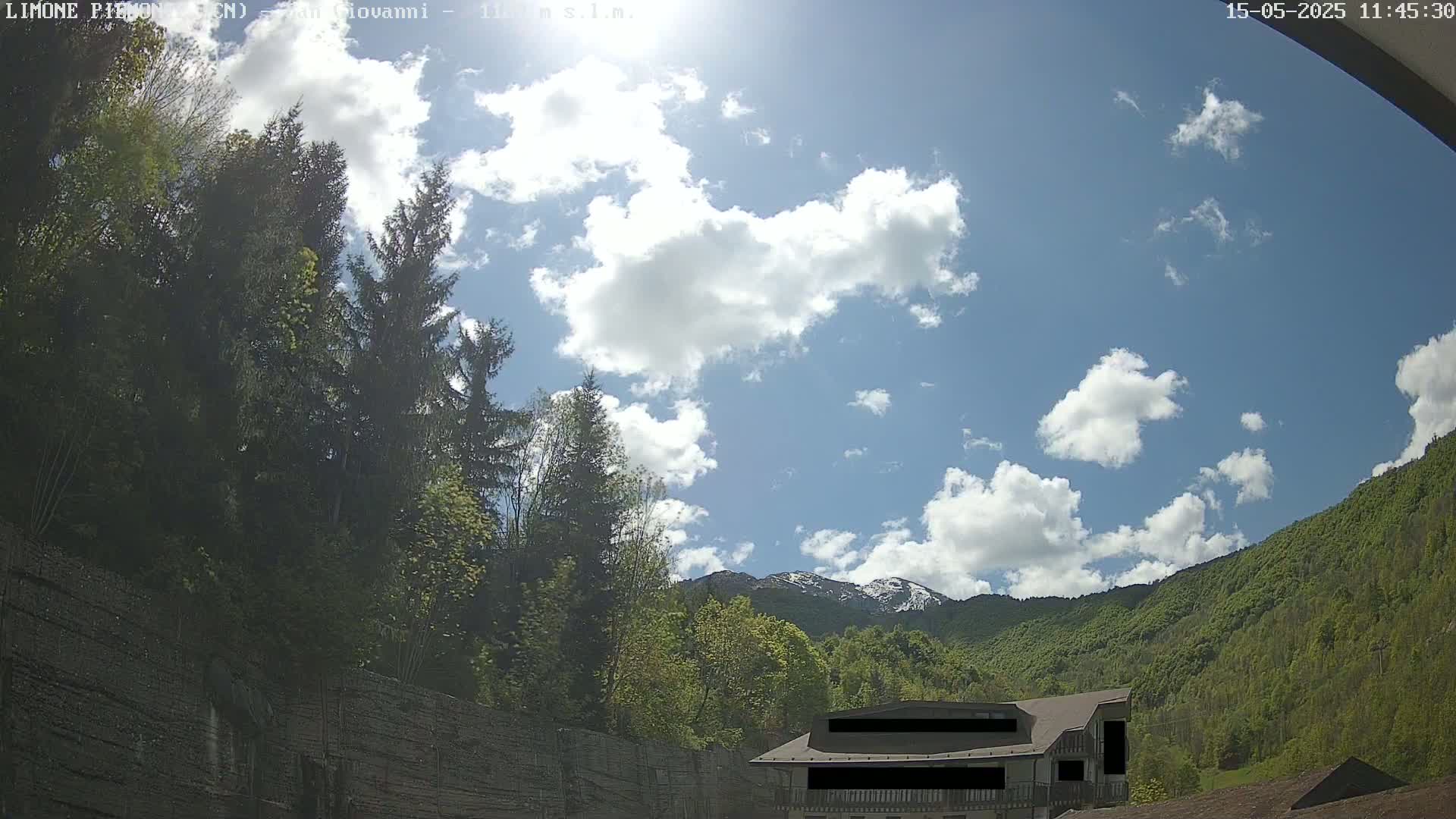 A partly sunny sky with fluffy white clouds overlooks a lush green mountainside, partially obscured by trees and a building.