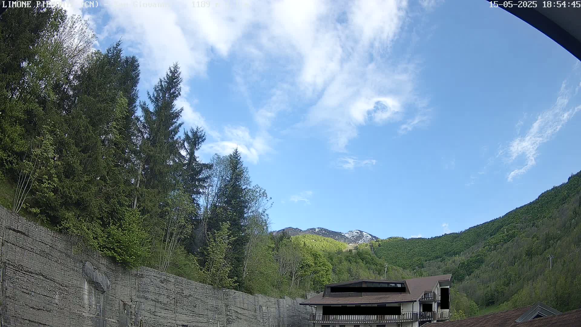 A partly cloudy sky overlooks a mountainous landscape with green trees and a building nestled among them, partially obscured by a tall retaining wall.