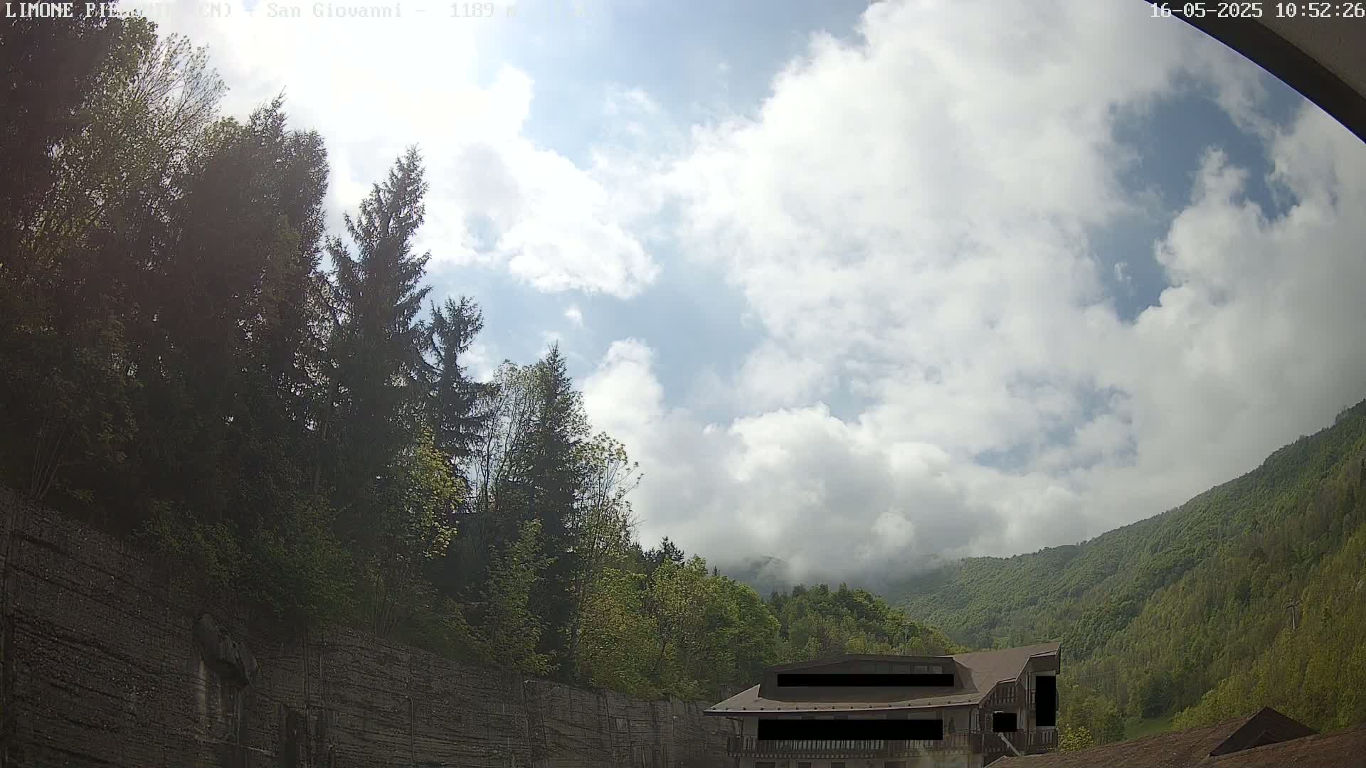 A partly cloudy sky is visible above a lush green mountainside with evergreens and a building partially obscured by a stone wall.