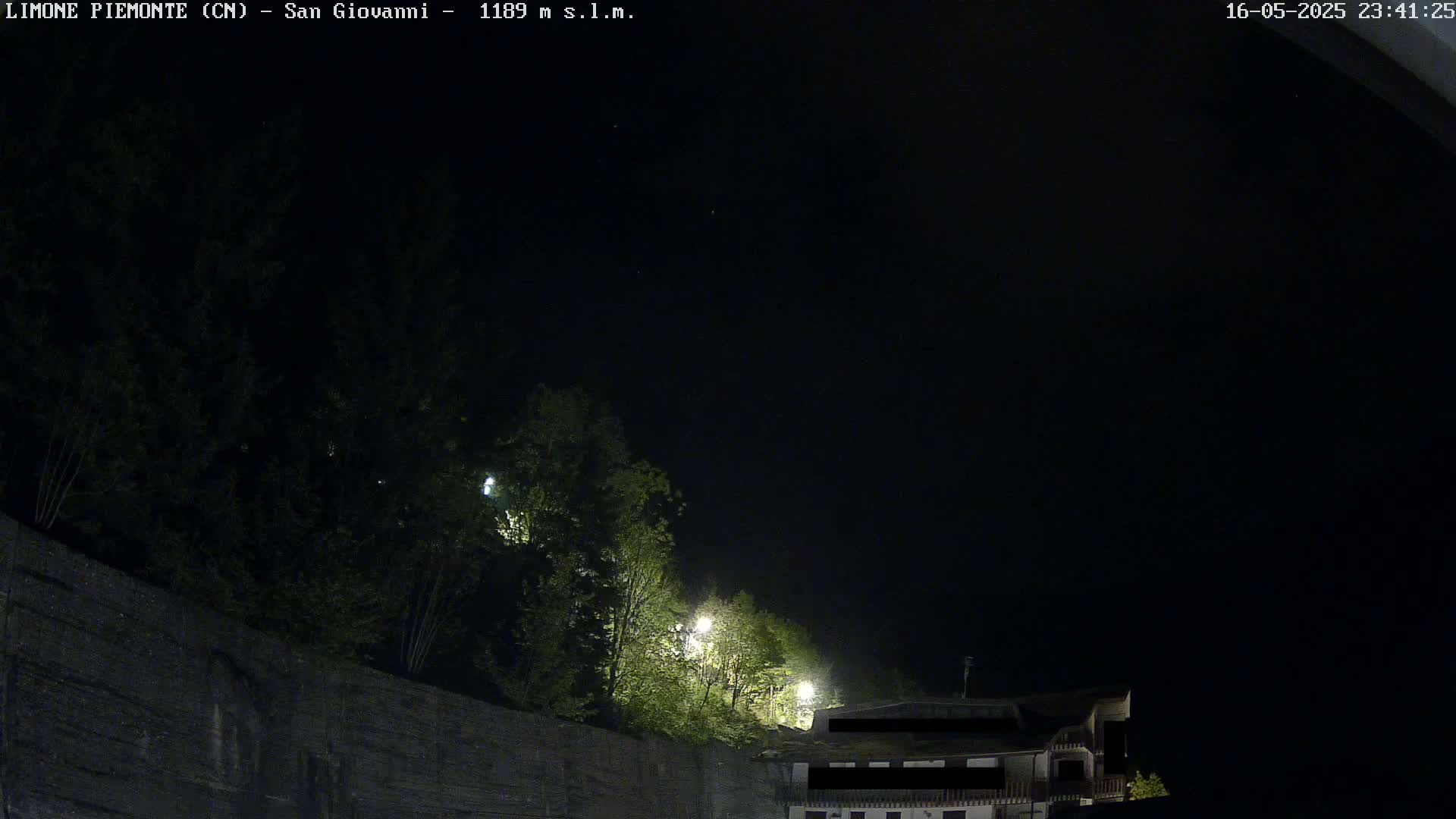 A nighttime view shows a dark sky over a stone wall topped with green trees and a building partially visible under some lights.