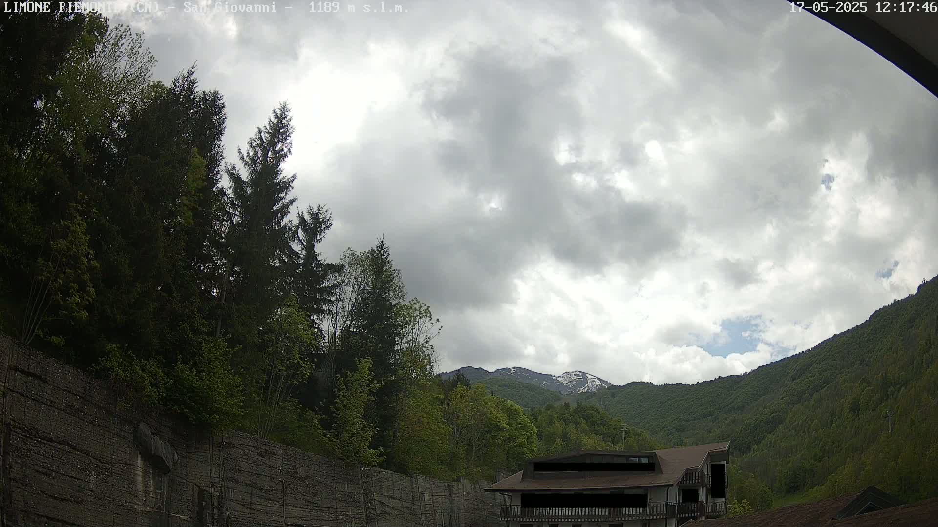 A partly cloudy sky overlooks a green mountainside with a building and trees, some snow visible on distant peaks.