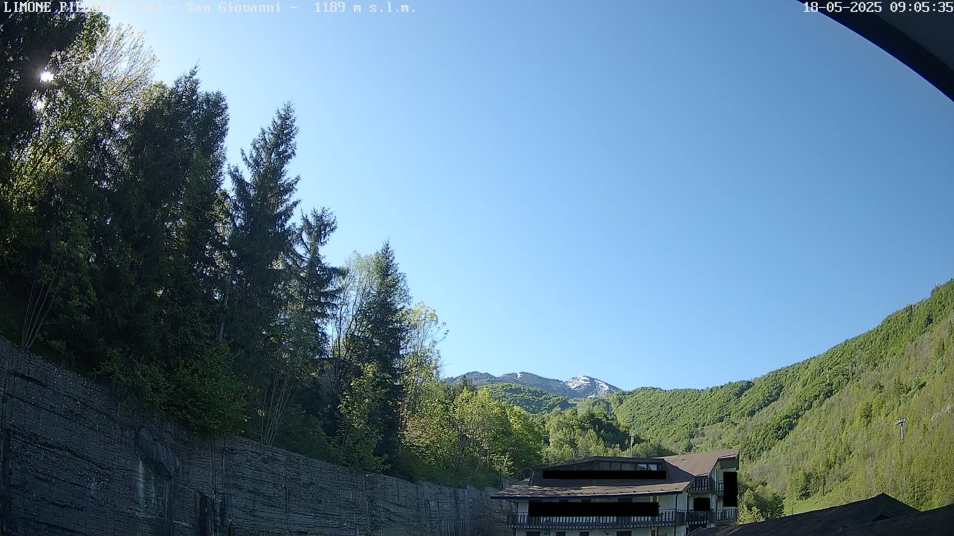 A sunny, clear day reveals a mountainside covered in lush green trees, partially obscured by a stone wall and evergreen trees in the foreground, with a building partially visible at the base.