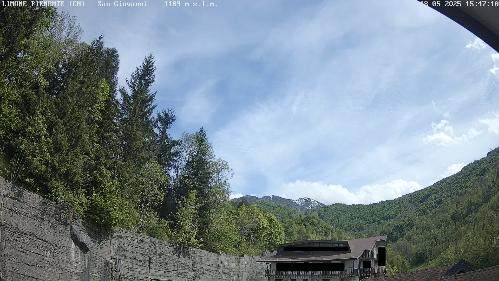 A partly cloudy sky is visible above a mountainous landscape with lush green trees and a building partially obscured by a concrete wall.