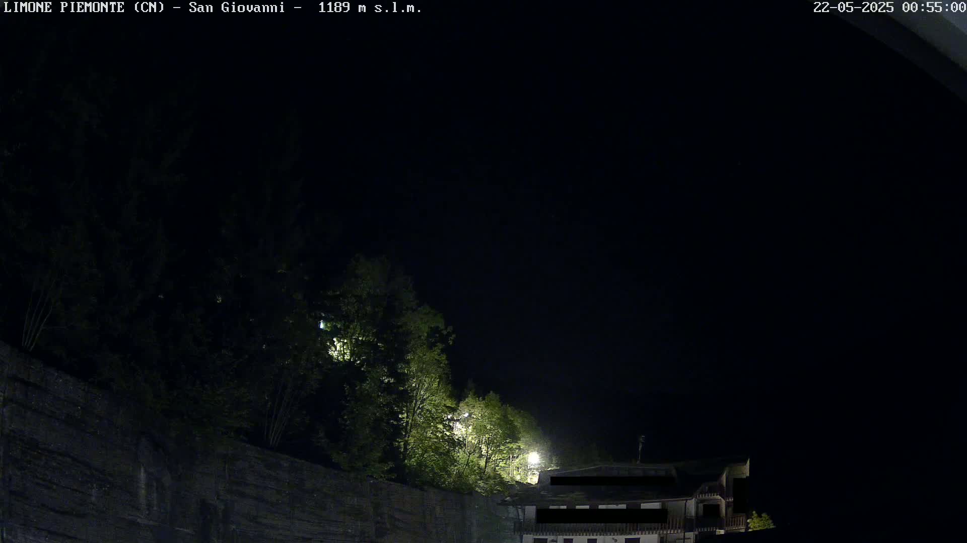 At night, a dark sky is above a stone wall behind which are some illuminated trees and a partially visible building.