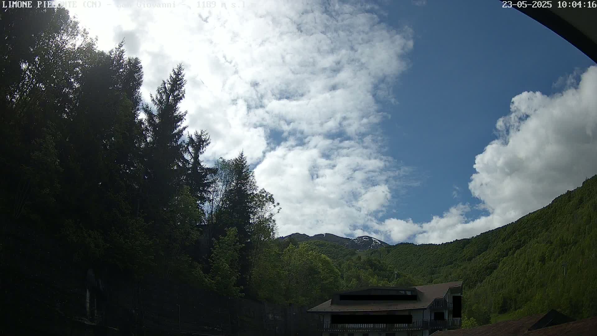 A partly cloudy sky over a mountain range partially covered in trees, with a building visible near the bottom right.
