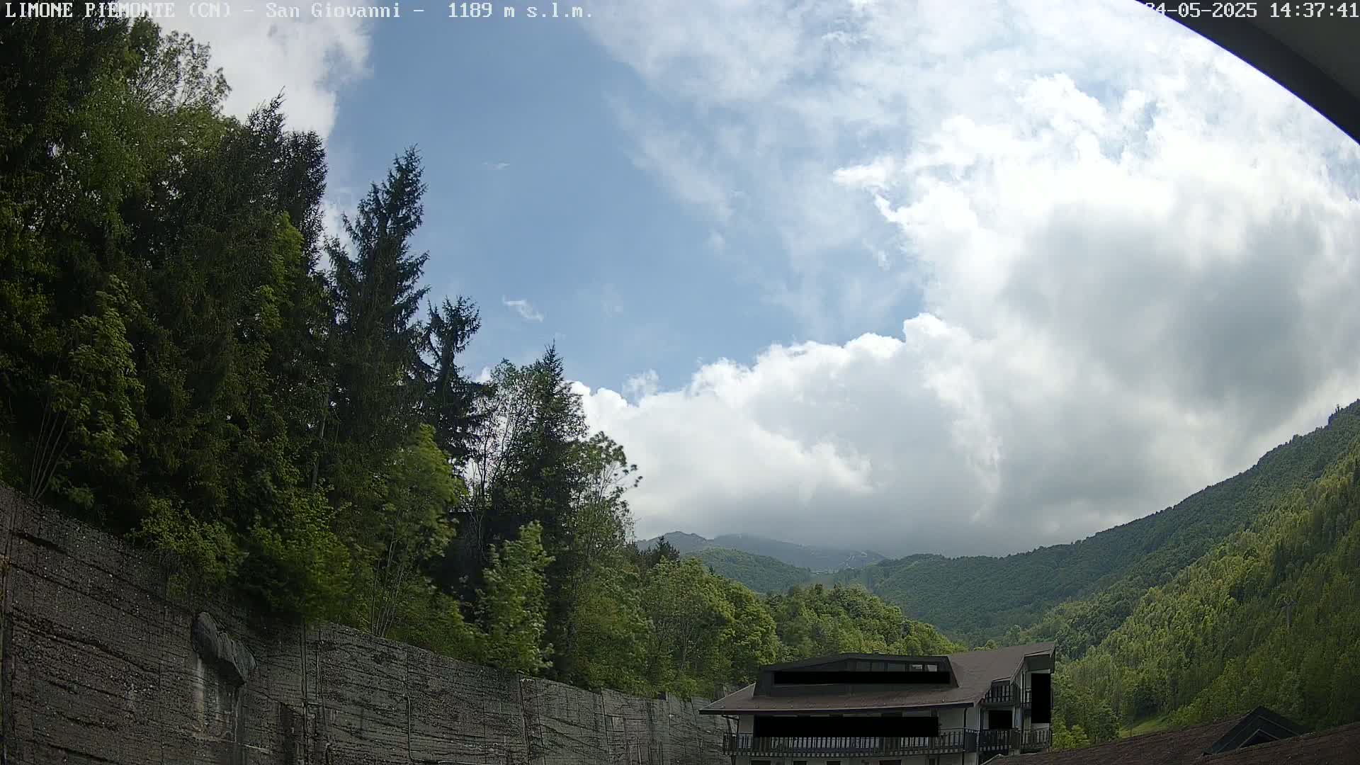 A partly cloudy sky over lush green mountains and trees, with a building partially visible in the foreground.
