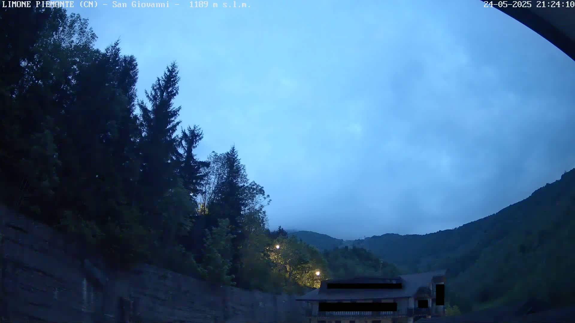 A nighttime view of a mountainside with dark green trees, a building partially visible, and a mostly clear, twilight sky.