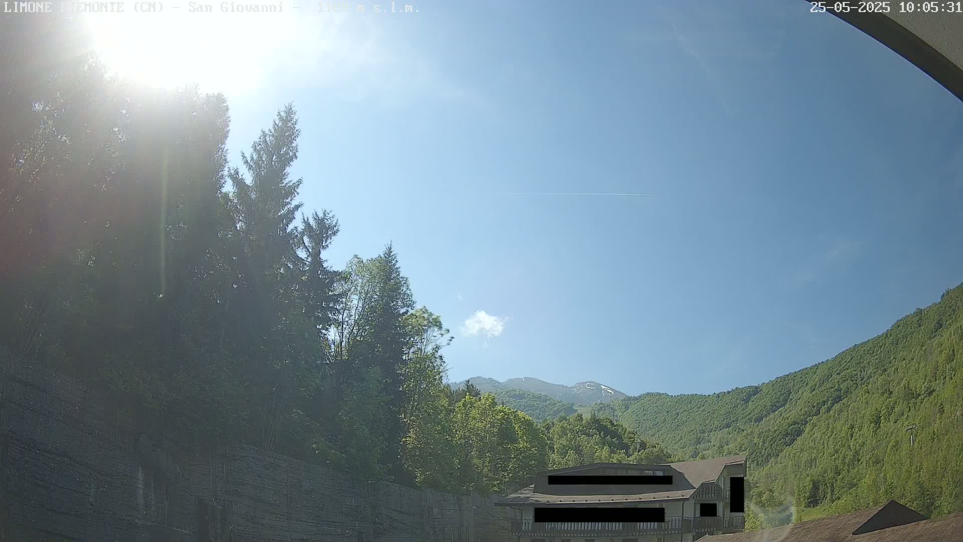 A sunny day with a mostly clear blue sky shows a mountainside covered in lush green trees, a building partially obscured, and several evergreen trees in the foreground.