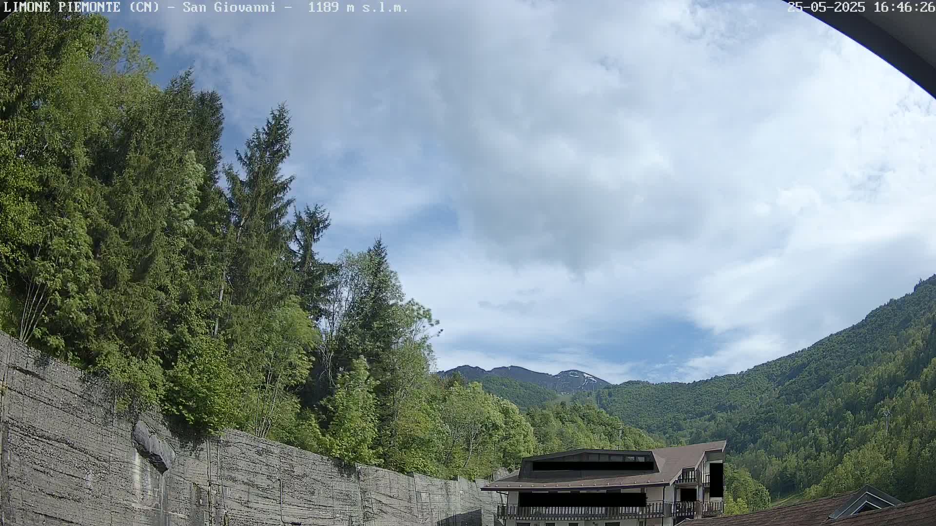 A partly cloudy sky overlooks a mountainous landscape of green trees and a building partially obscured by a retaining wall.