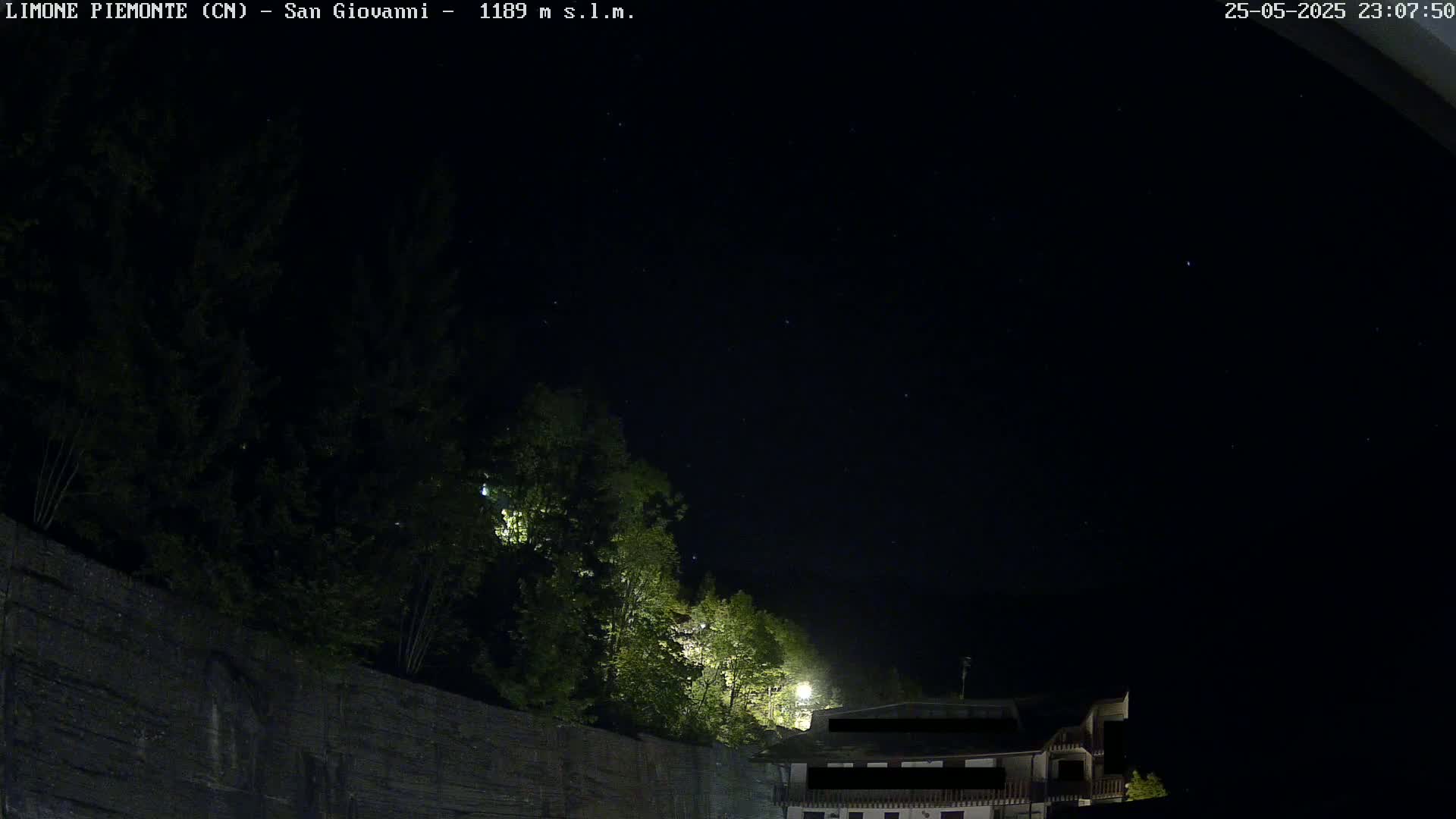 A nighttime view of a dark sky speckled with stars, overlooking a stone wall topped with dark green trees and a partially illuminated building.