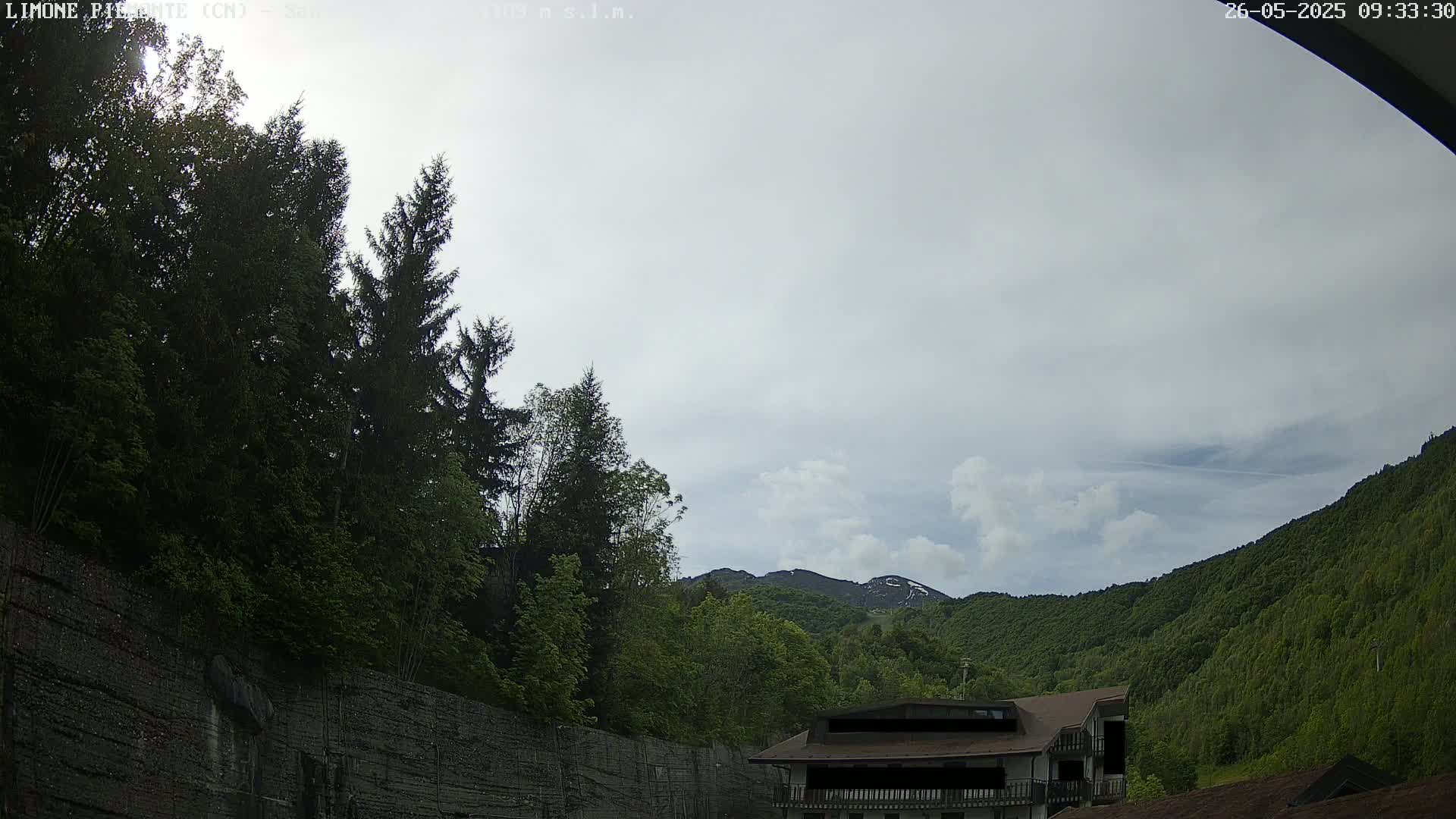 A mostly cloudy sky overlooks a mountainside covered in lush green trees and a building partially obscured by foliage.