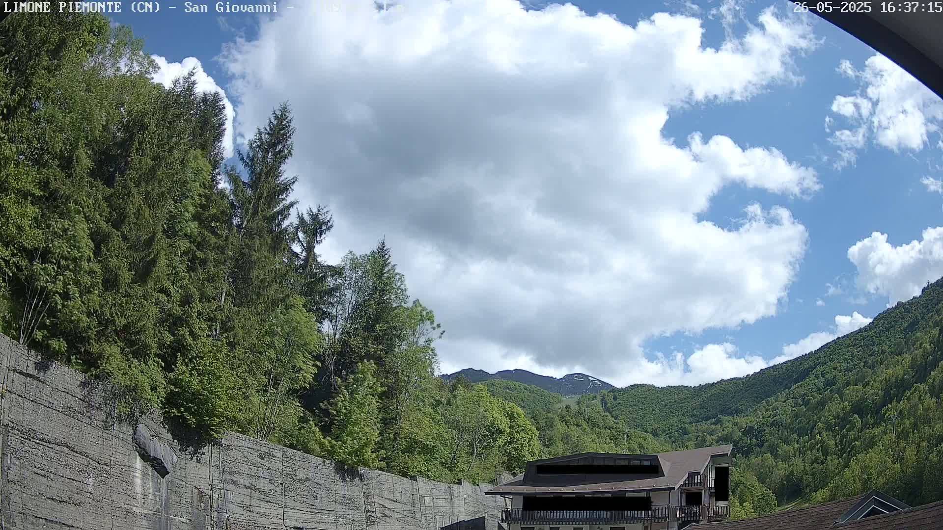 A partly cloudy sky is visible above lush green mountains and trees, with a building partially obscured by foliage in the foreground.