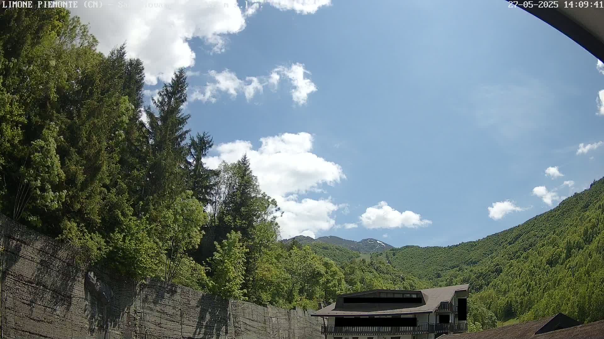 A partly cloudy sky overlooks lush green mountains and trees, with a building partially visible nestled among the greenery.