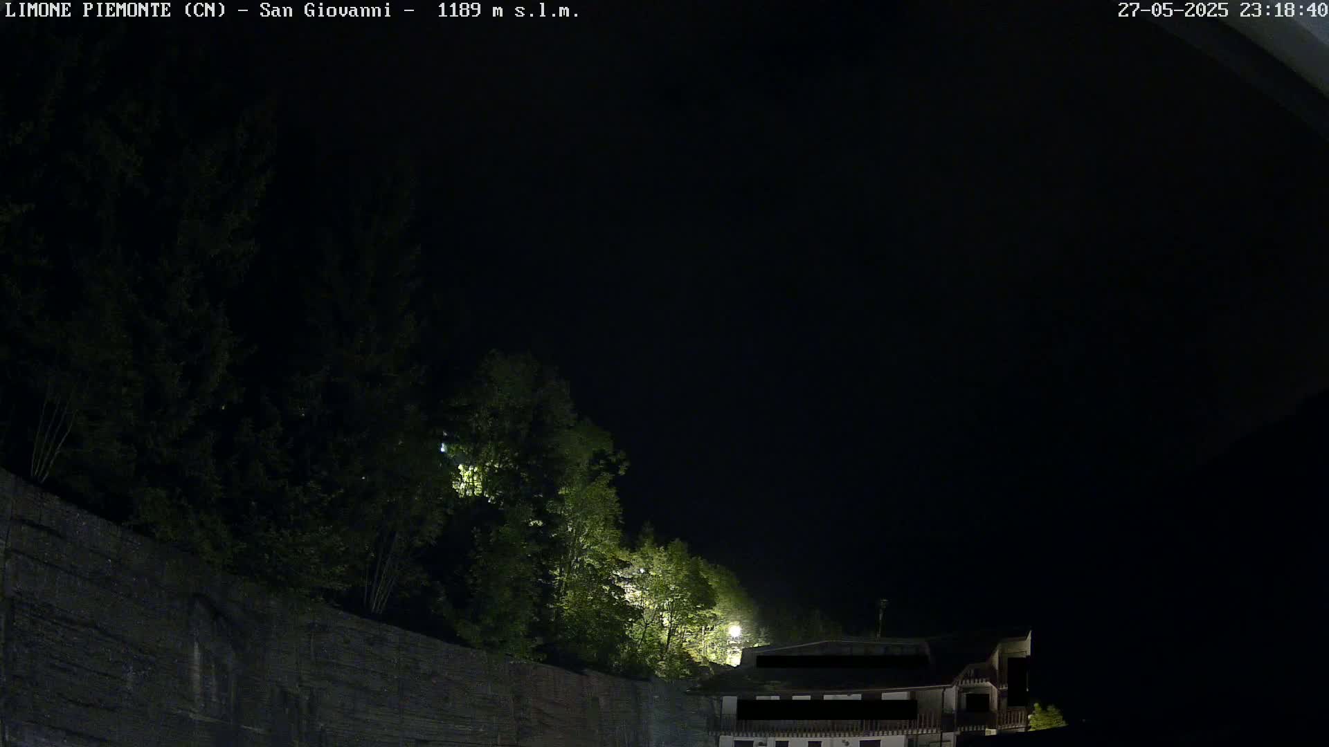 At night, a dark stone wall topped with illuminated trees sits in front of a dark building under a clear, black sky.