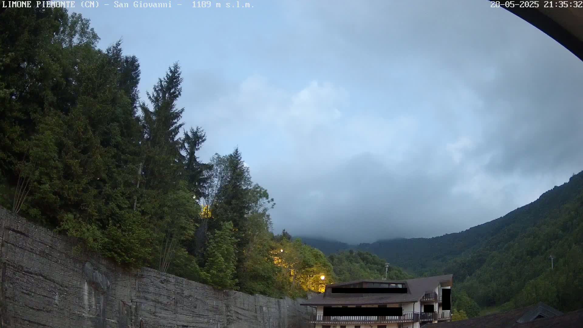A partly cloudy evening sky overlooks a mountainside with lush green trees and a building partially obscured by darkness.