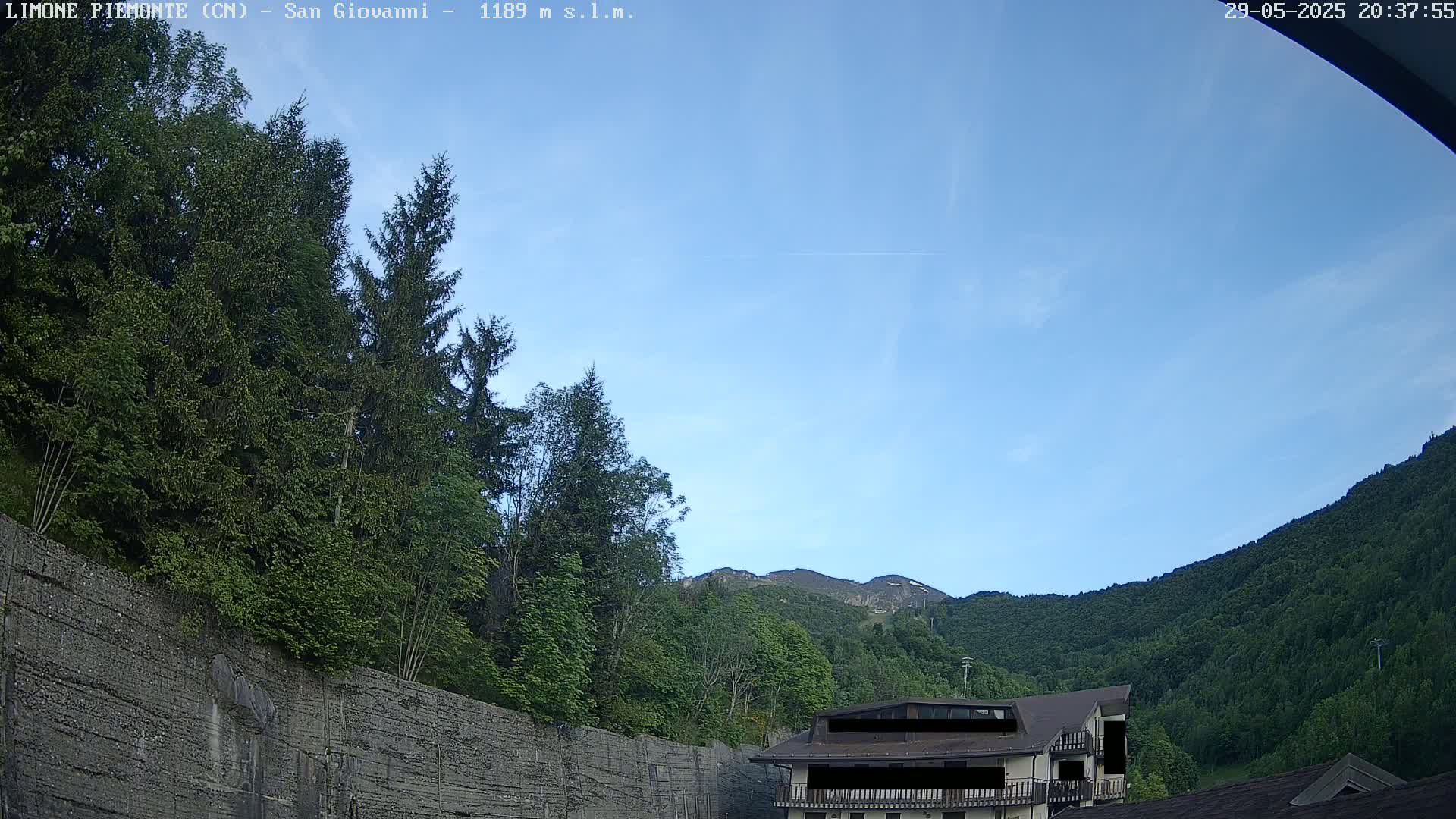 A mostly clear blue sky is visible above a mountainside covered in lush green trees and a building partially obscured by trees and a retaining wall.