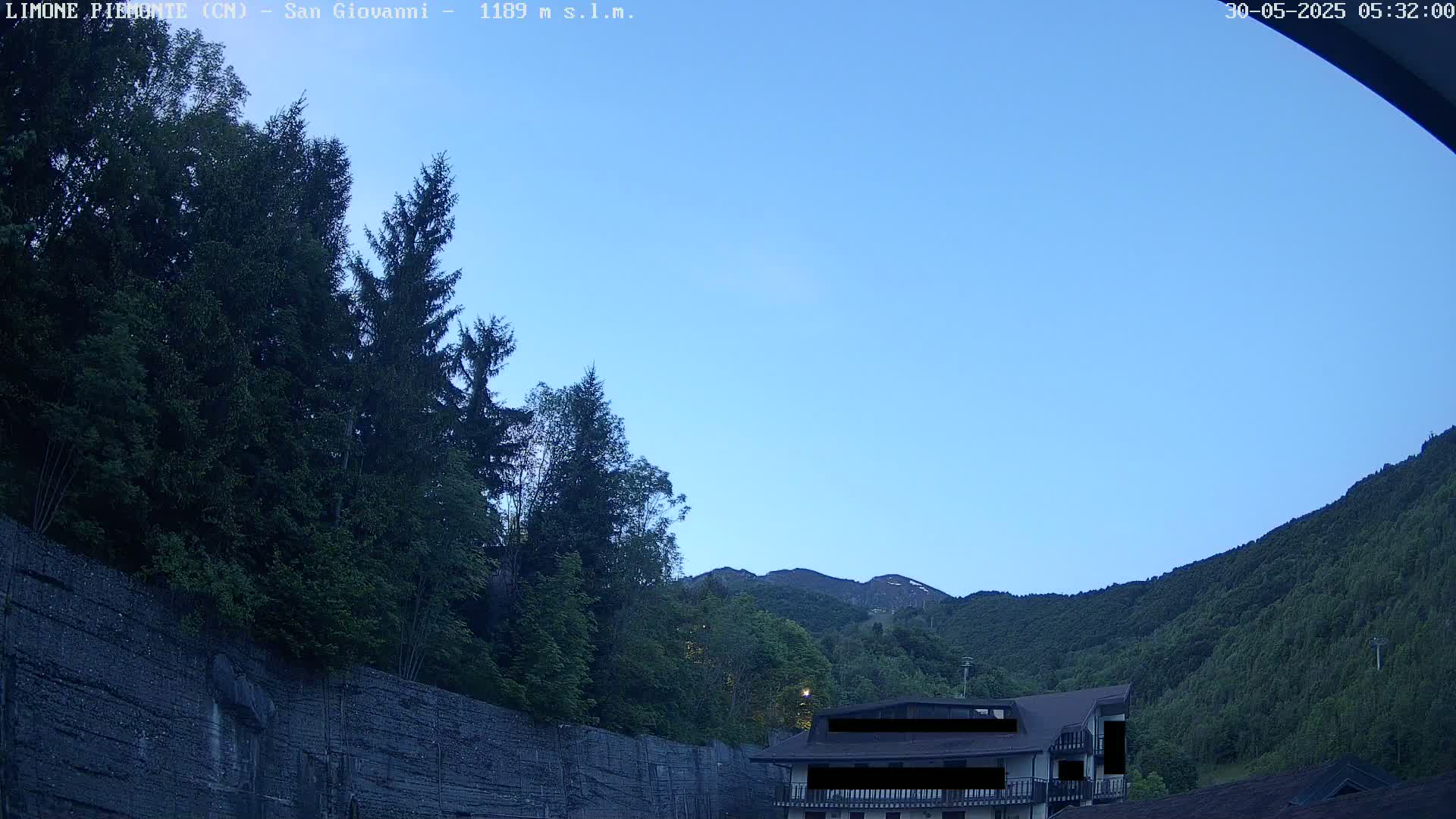 A clear blue sky overlooks a mountainside covered in lush green trees and a building partially obscured by foliage.
