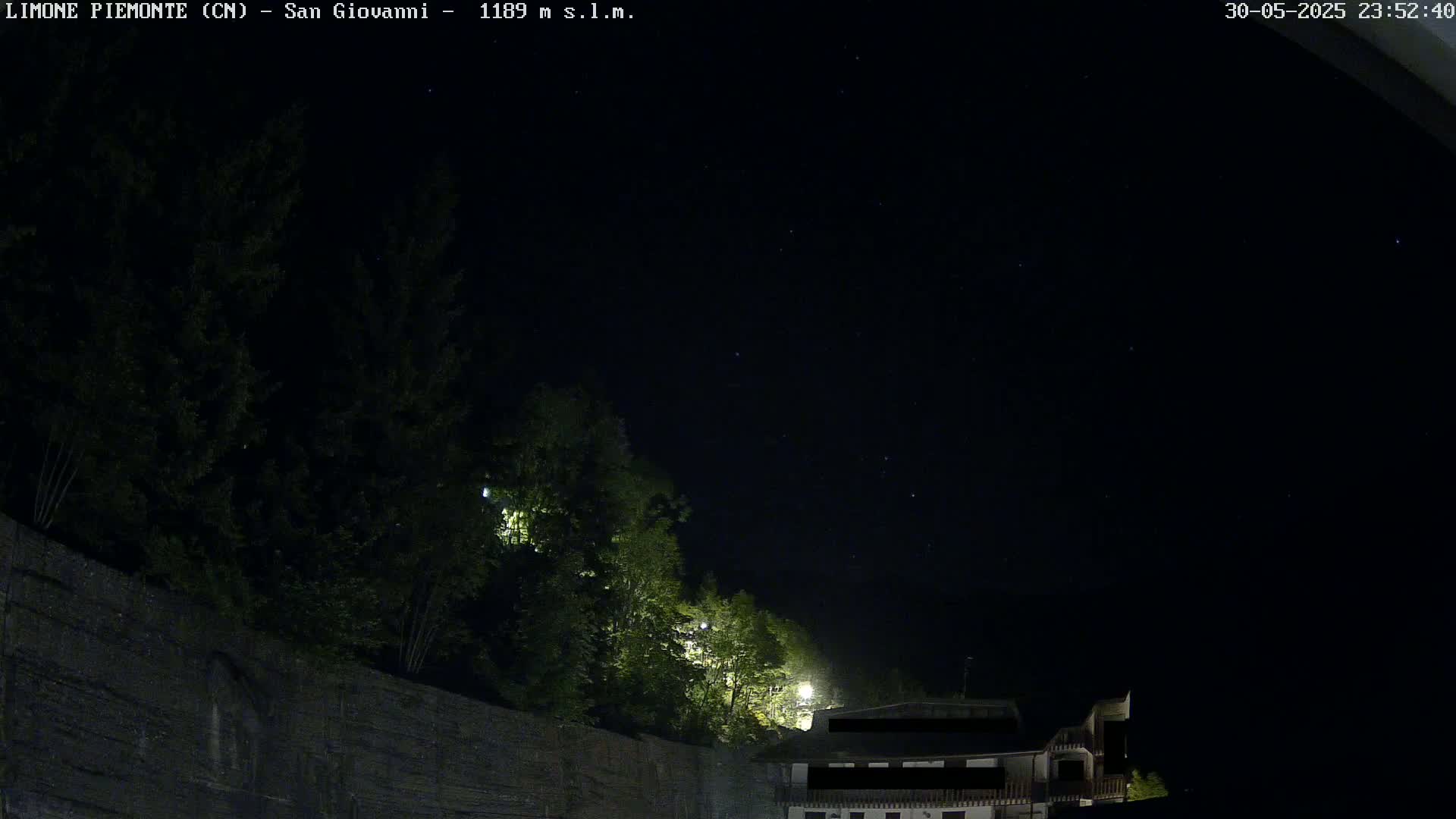 A nighttime view of a dark sky with stars visible above a stone wall and trees, partially illuminated by artificial light.