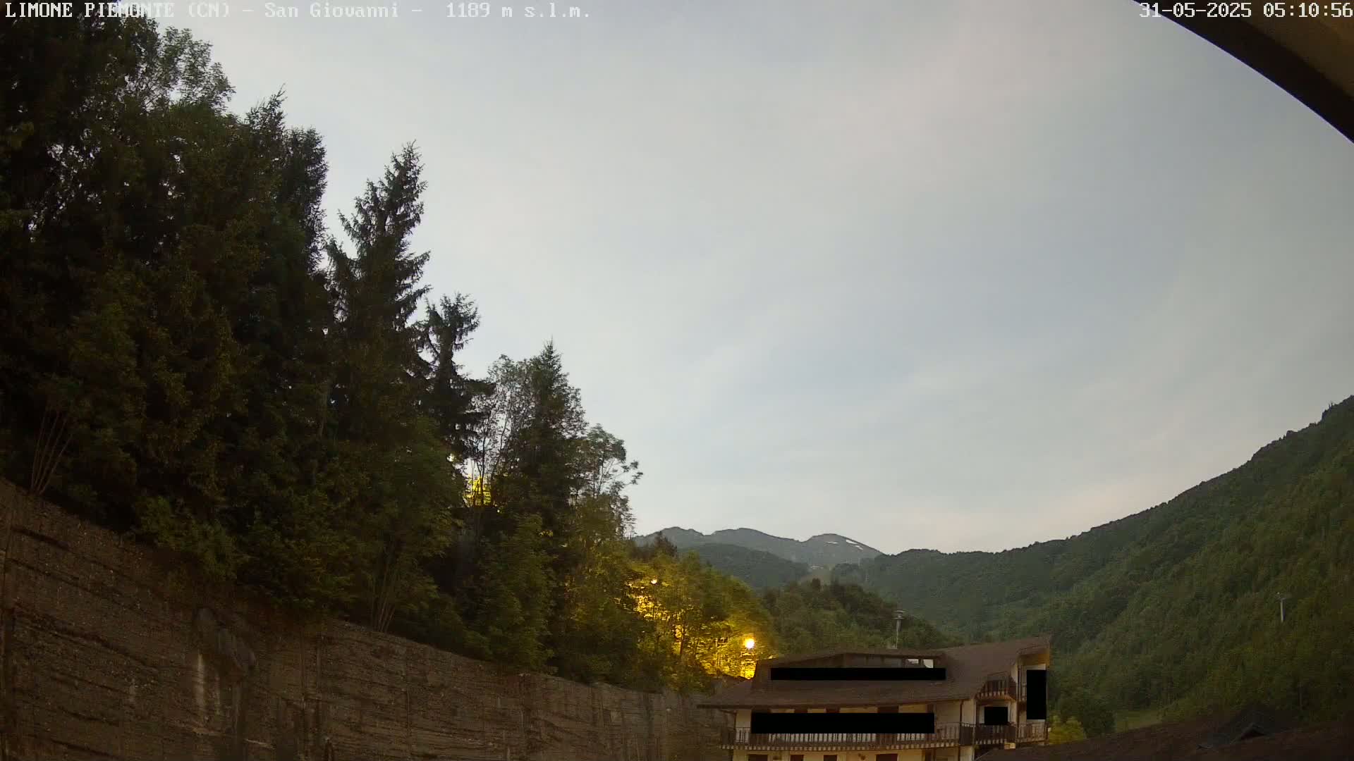 A mostly cloudy twilight sky overlooks a mountainous landscape with dark green trees, a stone wall, and a building partially visible in the lower right corner.