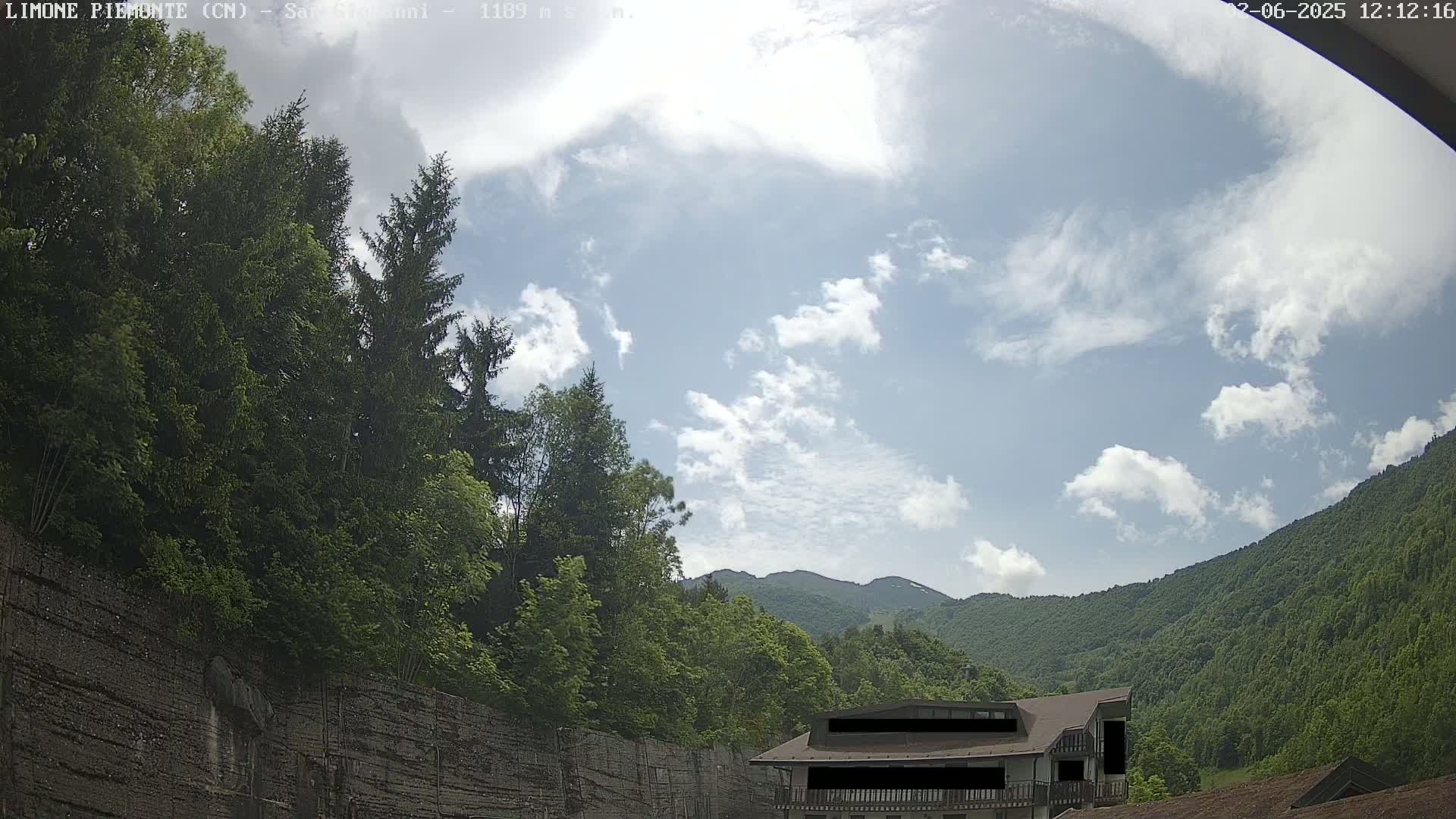 A partly cloudy sky overlooks a mountainous landscape with lush green trees and a building nestled among them.