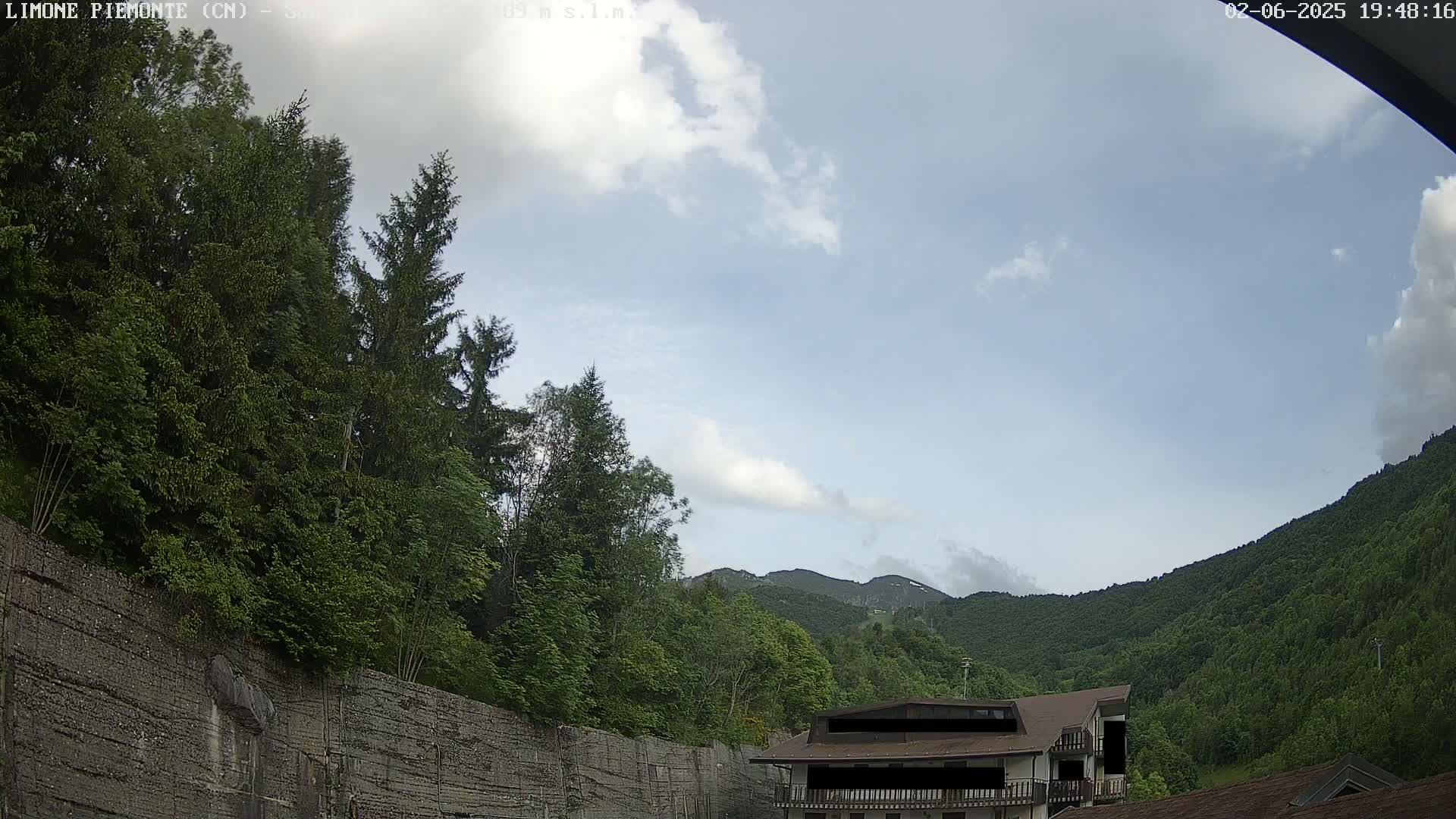 A partly cloudy sky overlooks a lush green mountainside with a building partially visible near a stone wall and dense trees.