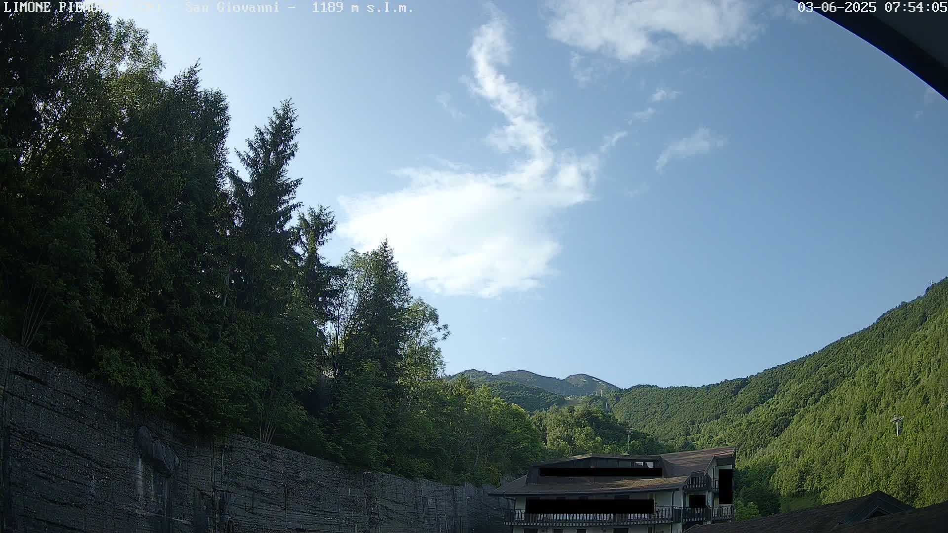 A partly cloudy sky overlooks a mountainside covered in lush green trees, with a building partially visible near a dark retaining wall.