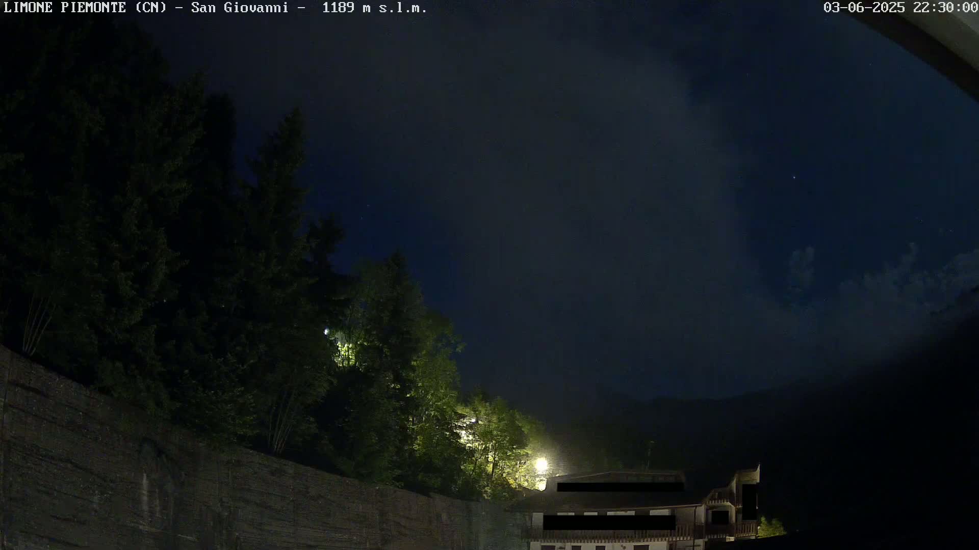 A nighttime view of dark green trees and a partially visible building under a mostly cloudy, dark blue sky.