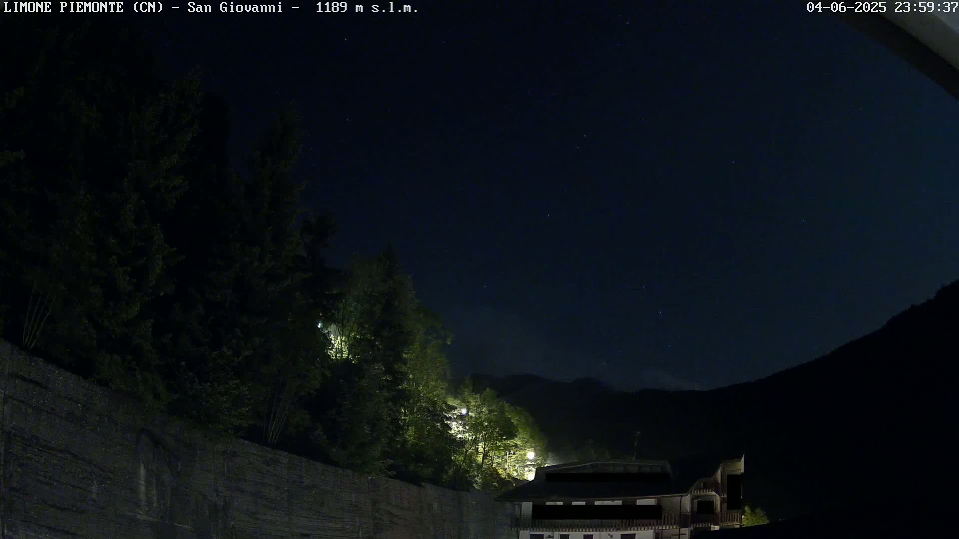 A night scene shows a dark mountainside under a clear, starry sky, with illuminated trees and a building partially visible near a stone wall.