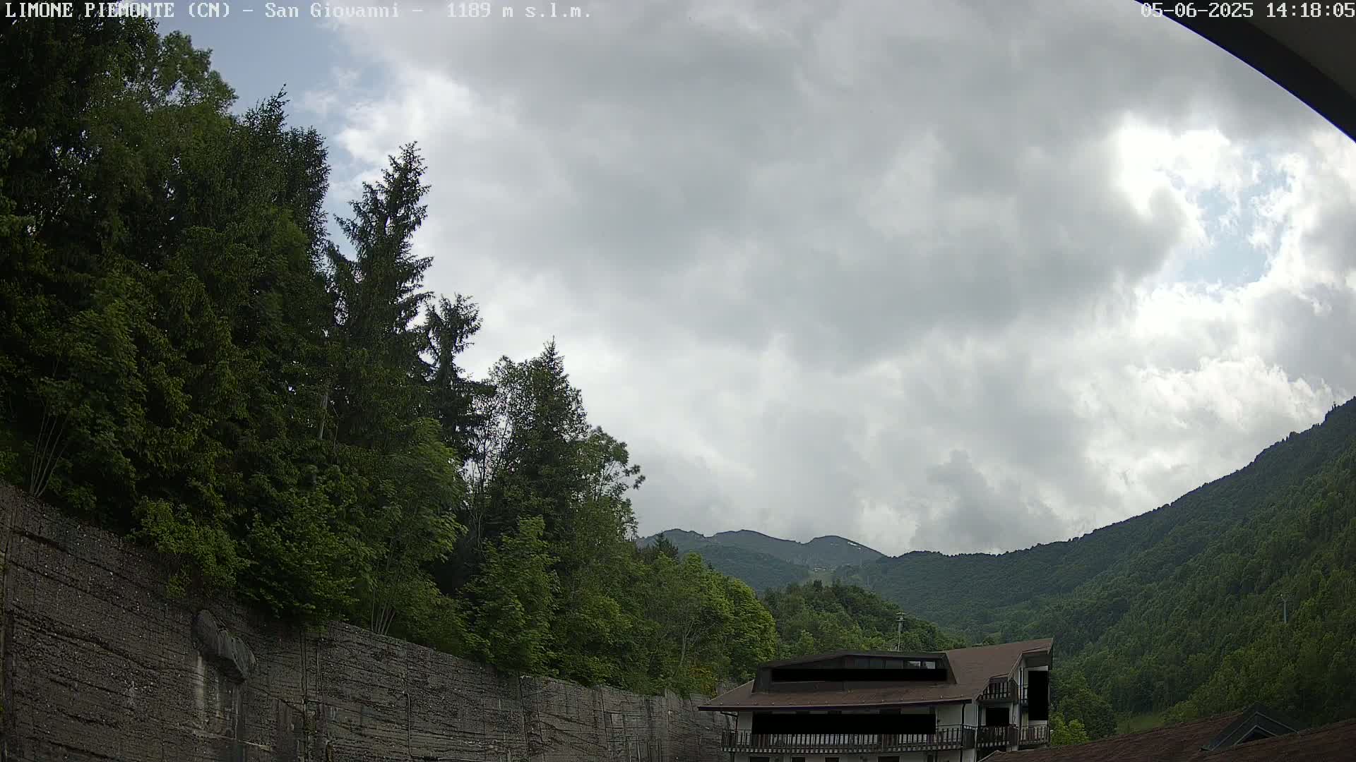 A mostly cloudy sky overlooks a mountainside covered in lush green trees and a building partially obscured by a wall.