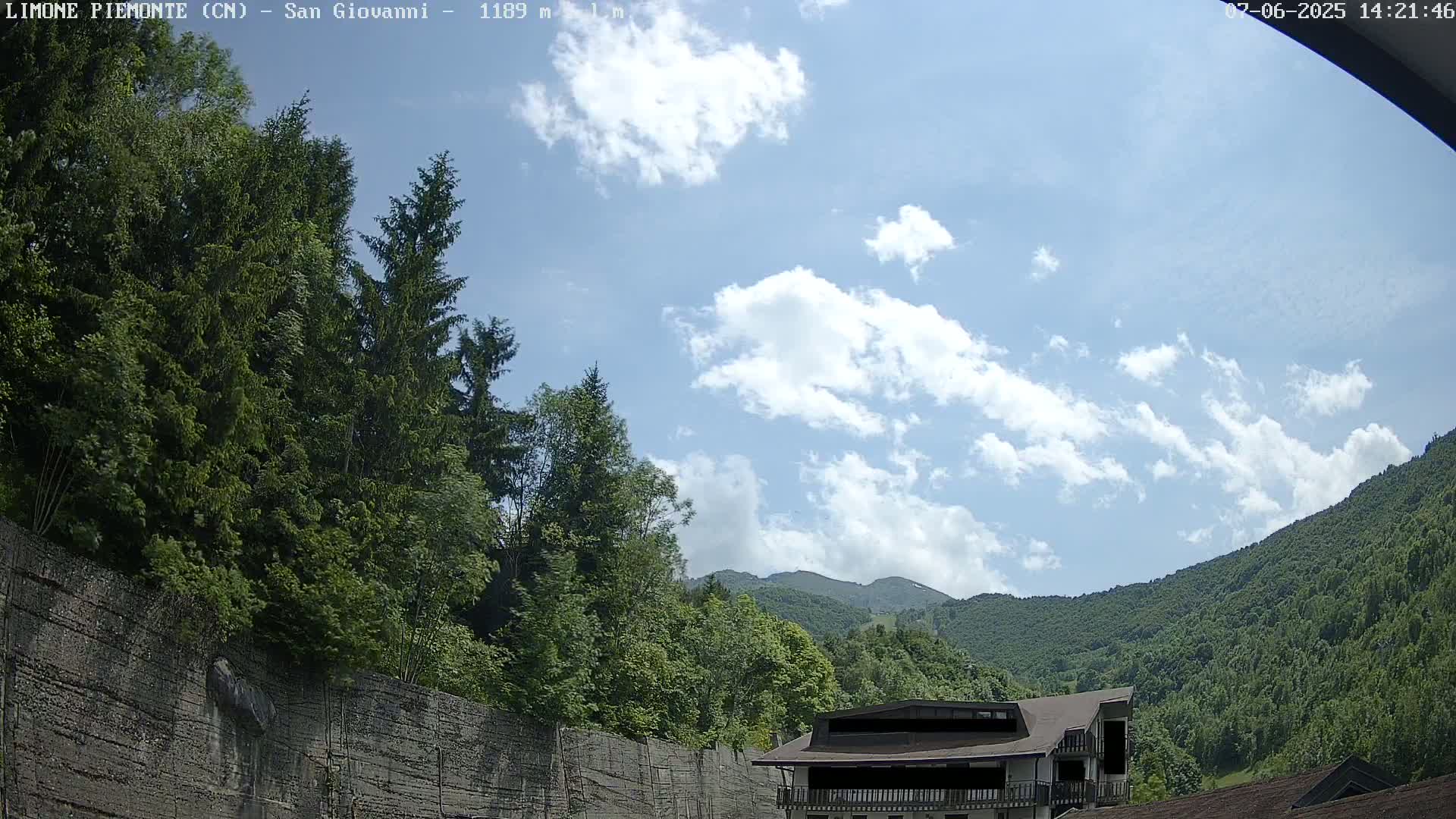A partly cloudy sky is visible above a lush green mountainside, partially obscured by a tall retaining wall and trees, with a building partially visible in the bottom right.