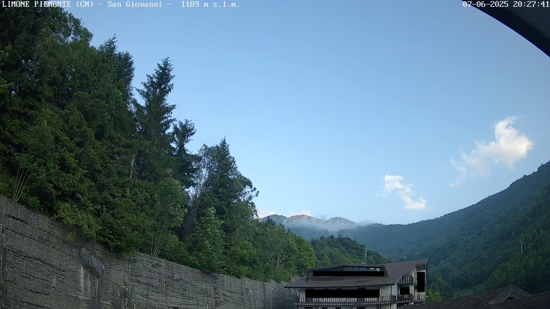 A mostly clear blue sky with a few small clouds overlooks a mountainside covered in lush green trees and a building partially obscured by trees.
