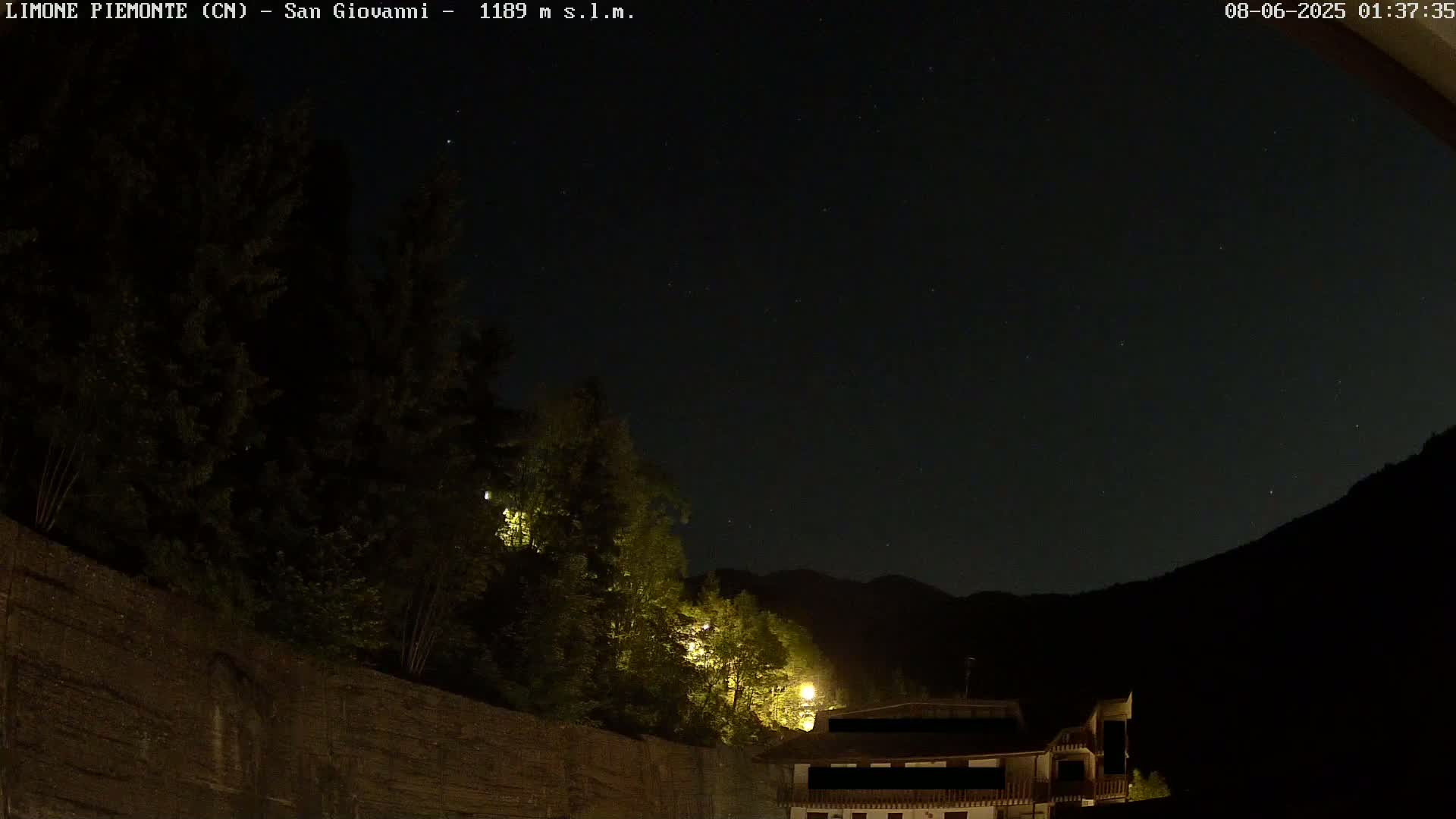 A nighttime view of a dark mountainside with a stone wall and trees illuminated by lights, under a clear, starry sky.