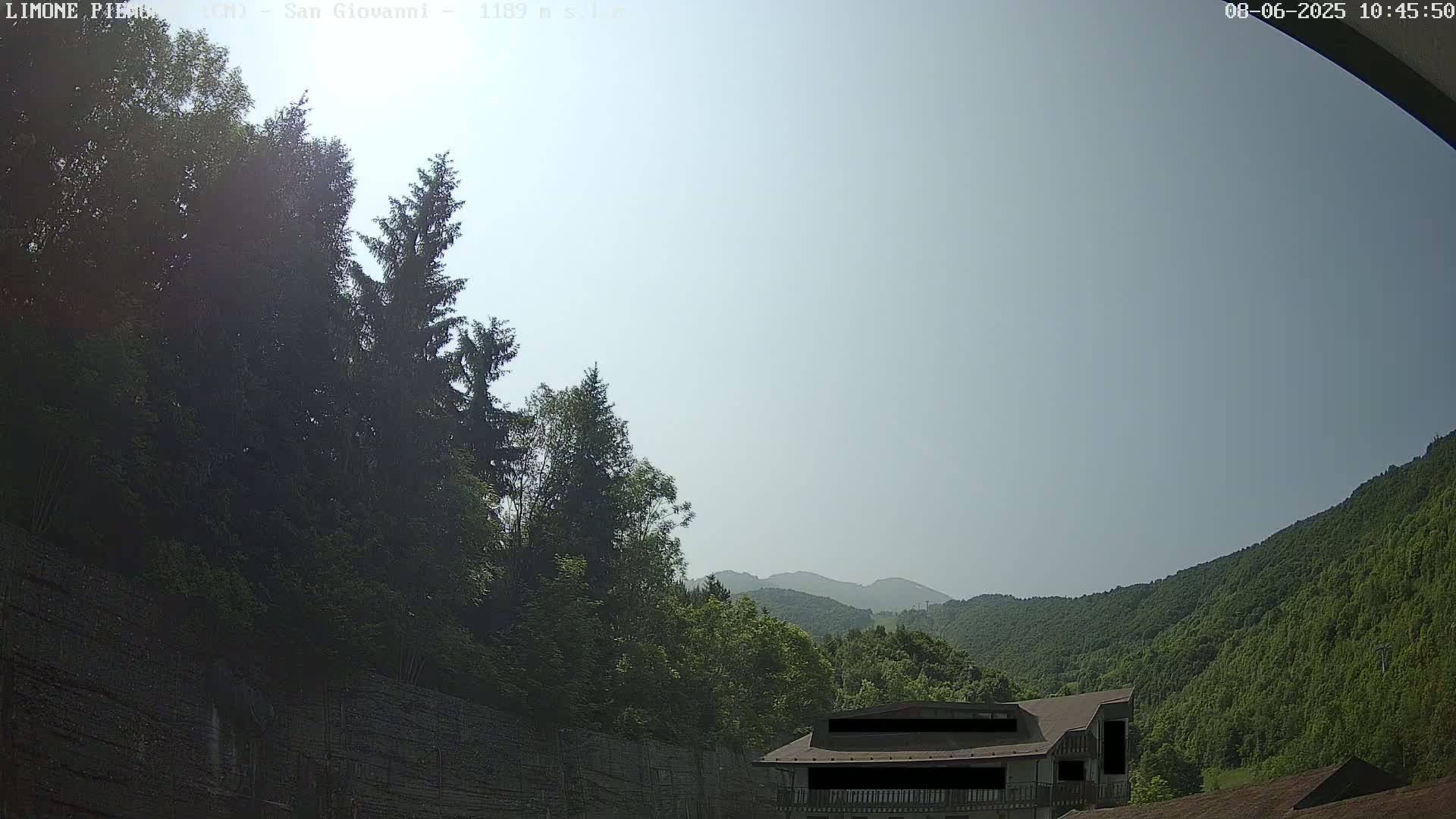 A hazy, sunny day reveals a mountainside covered in lush green trees, with a building partially visible near the bottom and taller trees in the foreground.