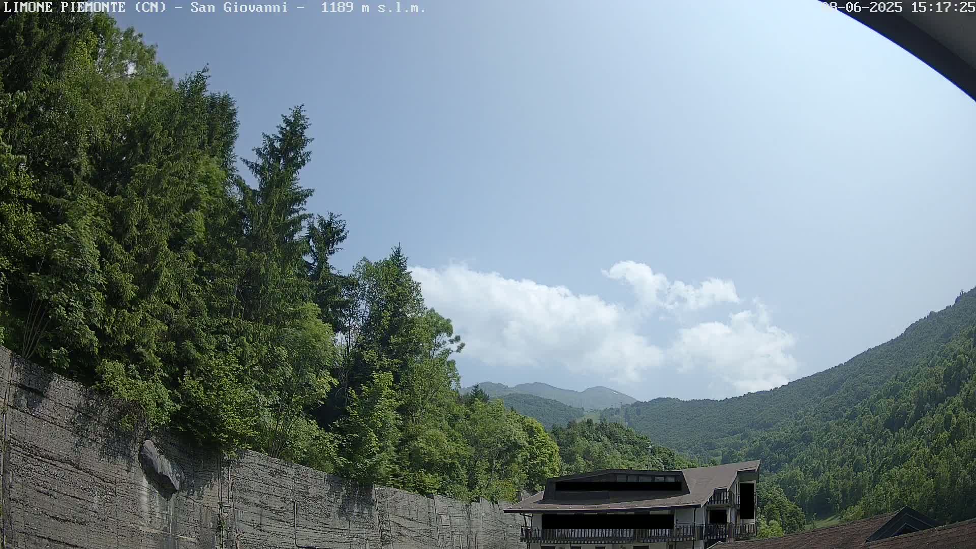 A partly cloudy sky overlooks a mountainous landscape with lush green trees, a concrete retaining wall, and a building partially visible.