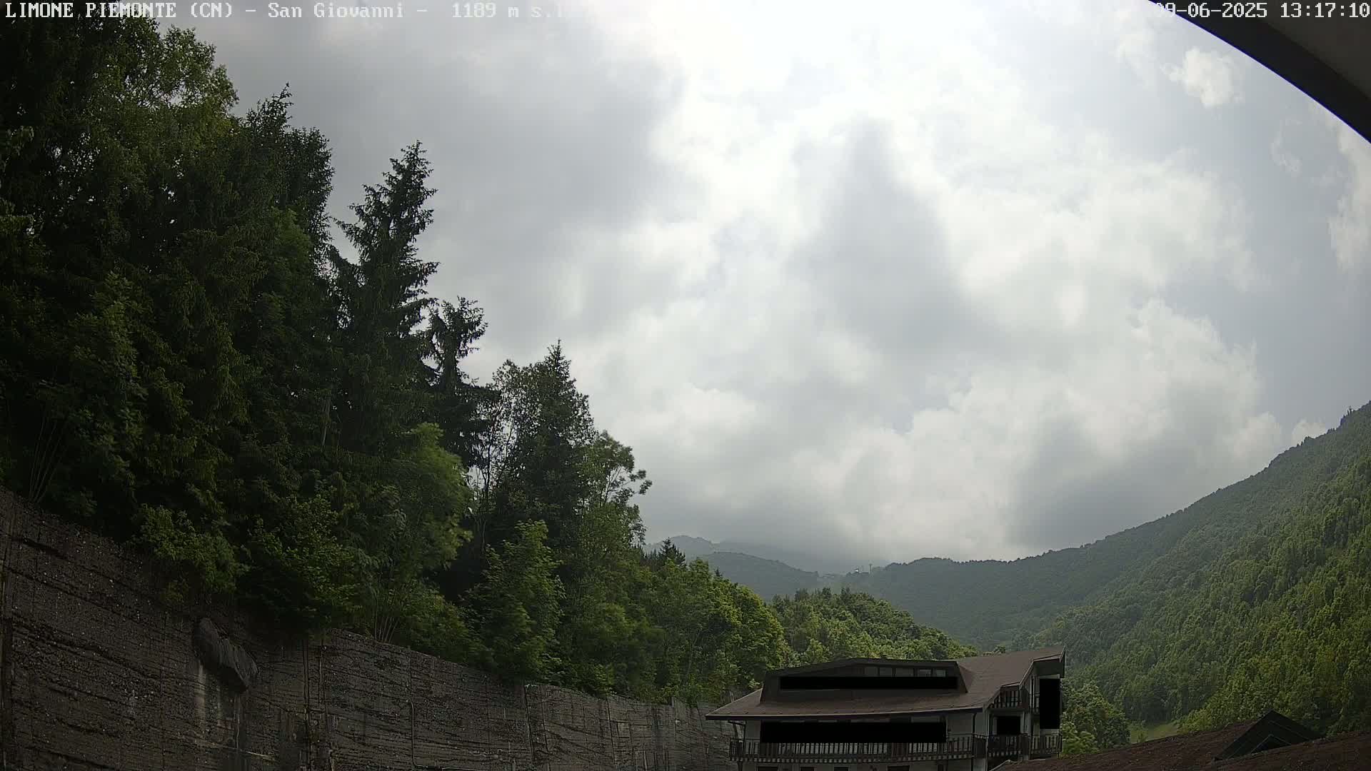 A cloudy day reveals a mountainside covered in lush green trees and a building nestled amongst them, near a high retaining wall.