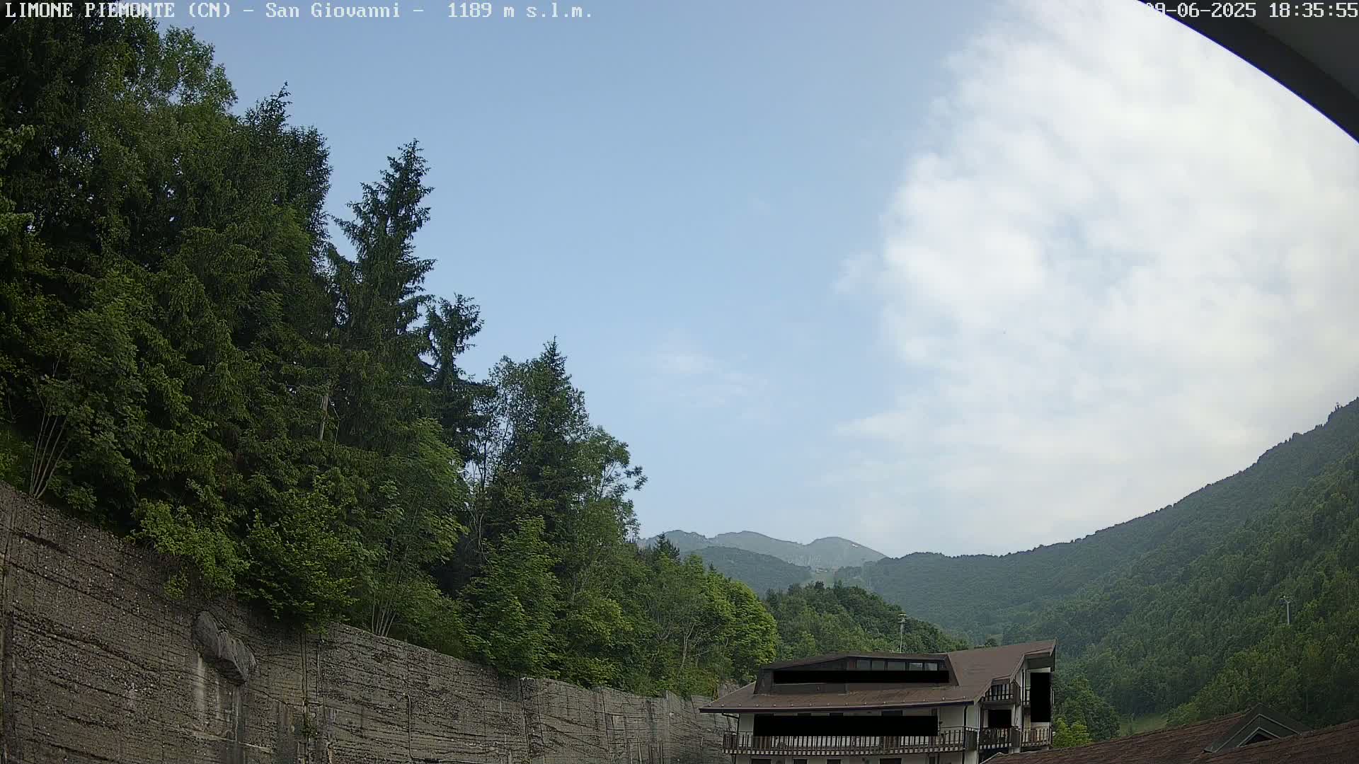 A partly cloudy sky overlooks a mountainside covered in lush green trees and a building partially obscured by the trees.