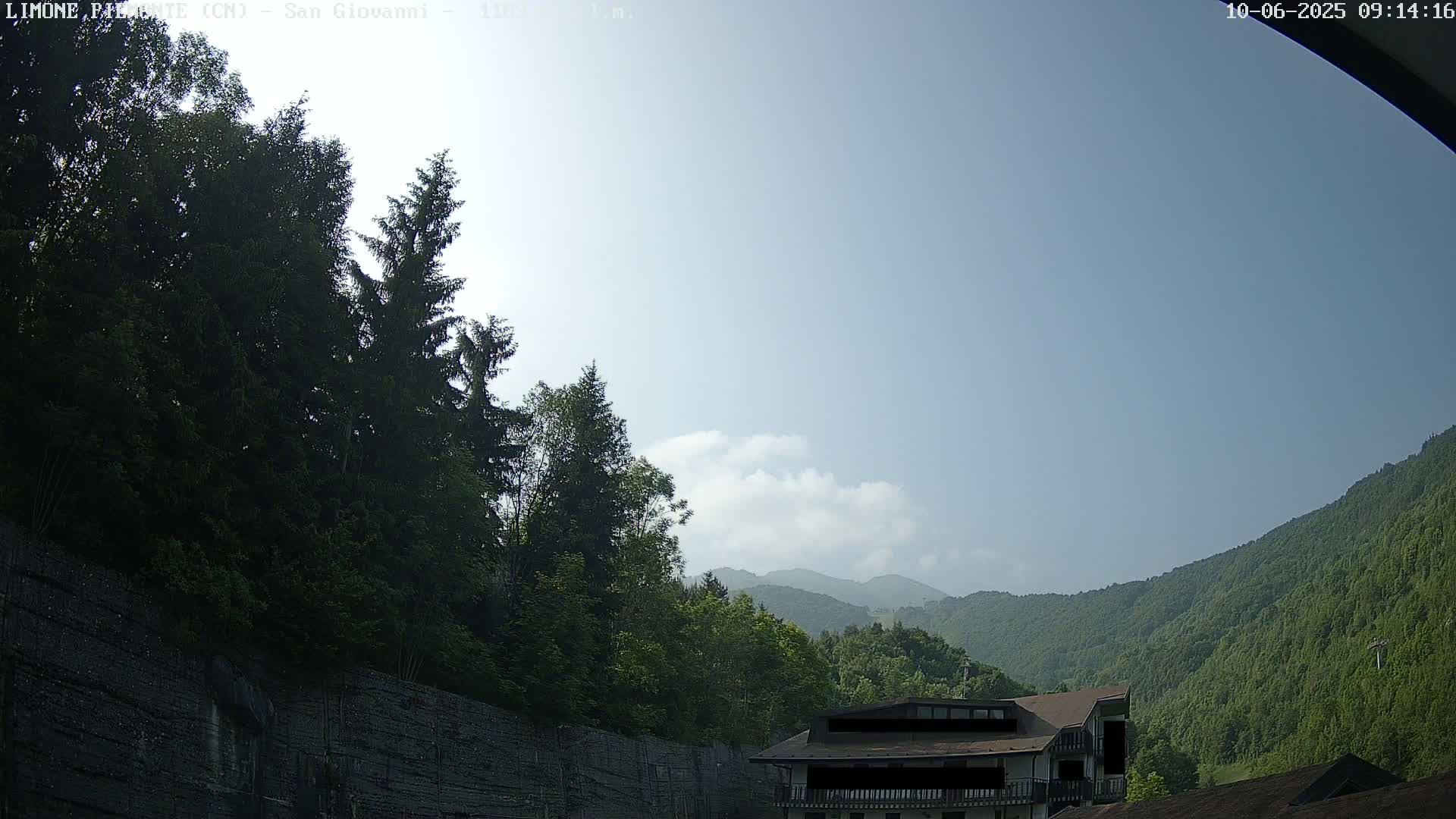 A partly cloudy sky over a mountainous landscape, with lush green trees and a building partially visible among them.