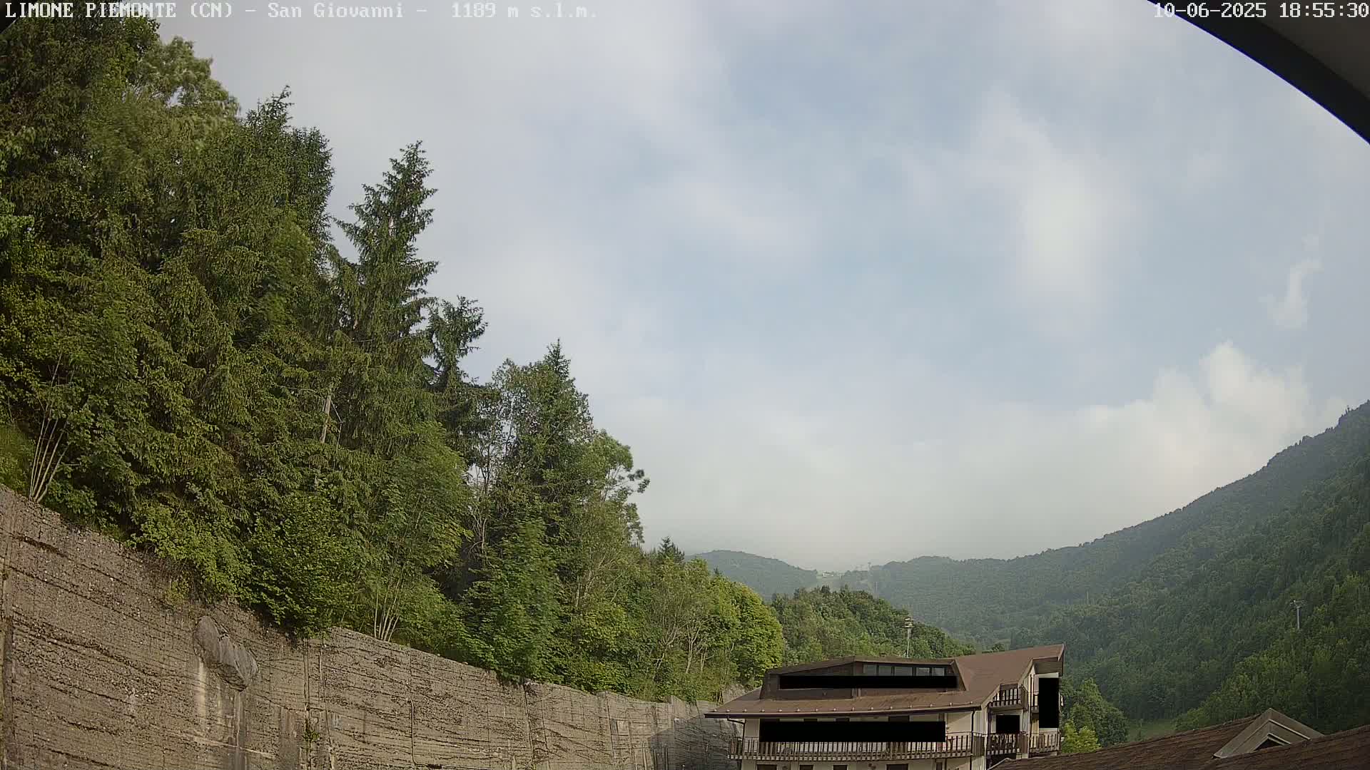 A partly cloudy sky overlooks a mountainous landscape with lush green trees and a building partially visible near a retaining wall.